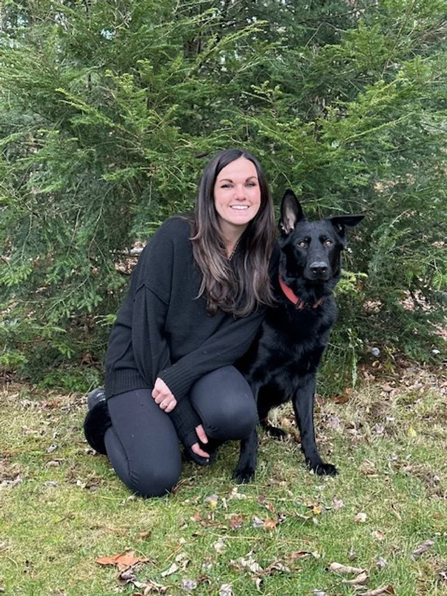A woman kneeling on the grass next to a black dog in front of green shrubbery.