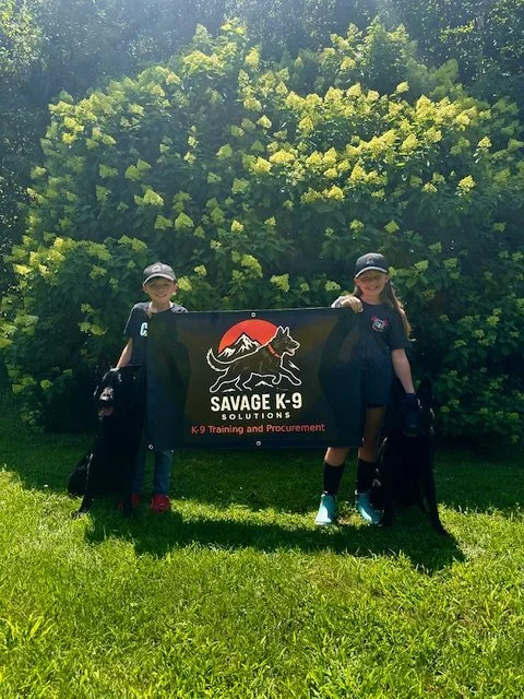 A black banner that reads "Savage K-9 Solutions, K-9 Training and Procurement." They are outdoors with a backdrop of lush green trees and yellow flowering bushes.
