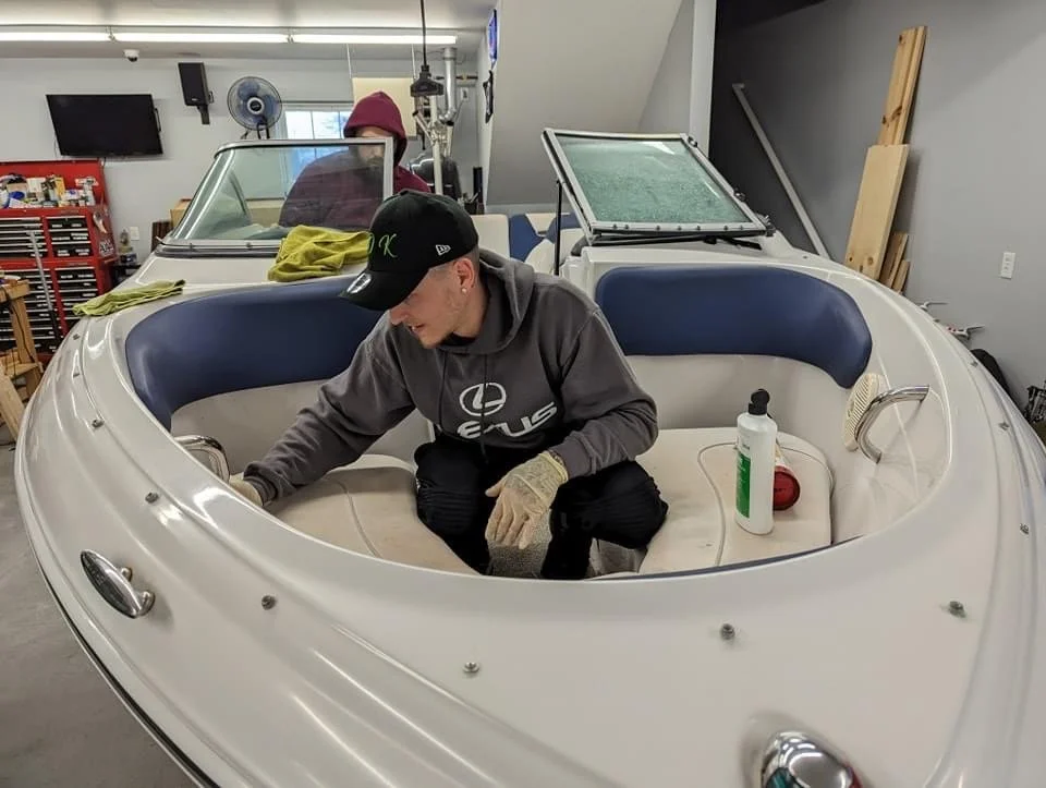 Person cleaning or maintaining a boat inside a workshop, wearing gloves and a baseball cap, with cleaning supplies nearby.