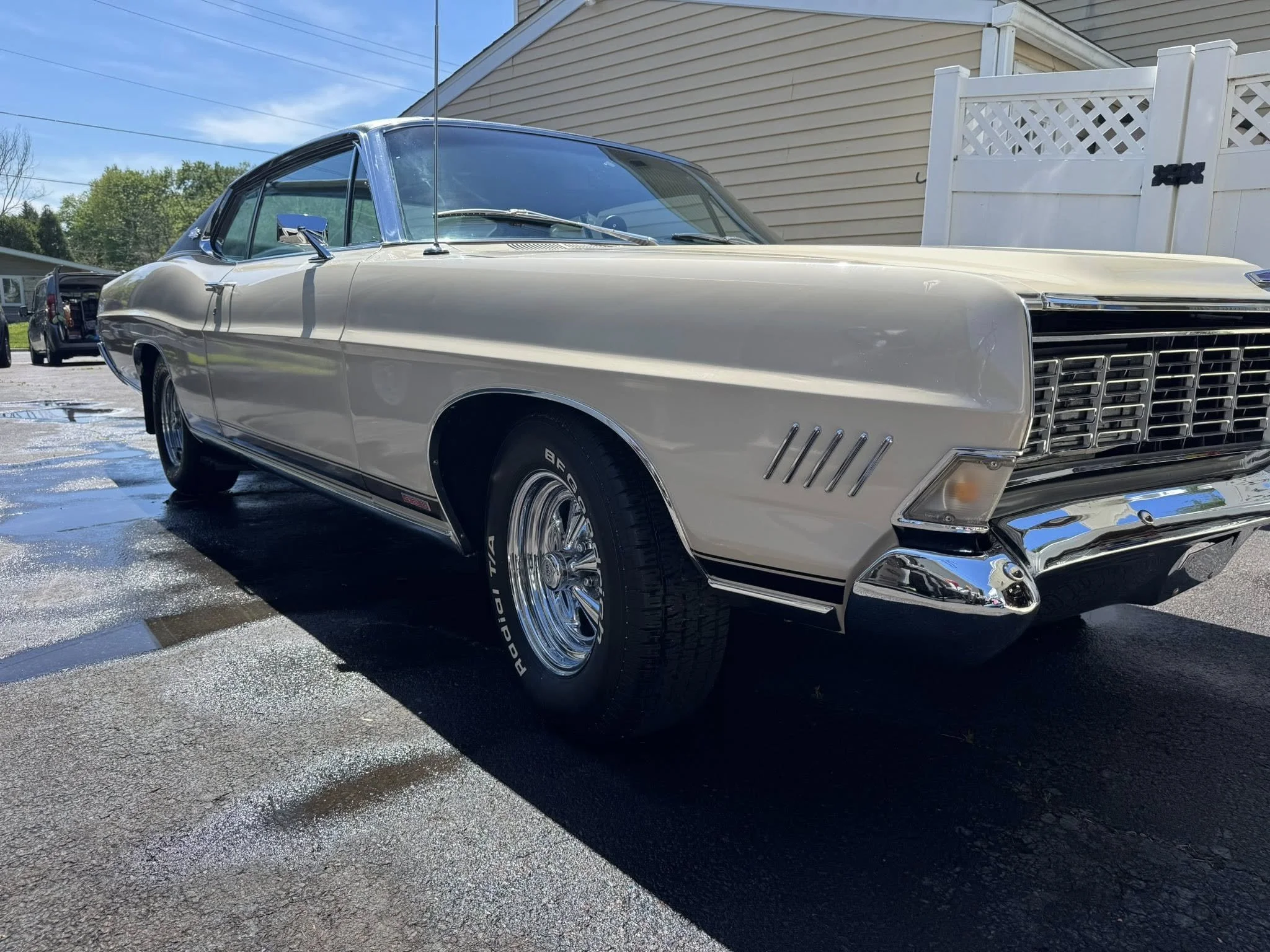 A vintage cream-colored car parked on a wet driveway next to a white fence with a house in the background.