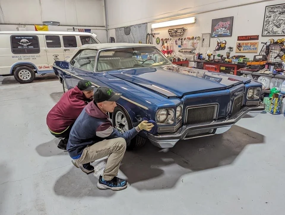 Two young men cleaning a vintage blue car inside a garage with tools and signs on the wall.