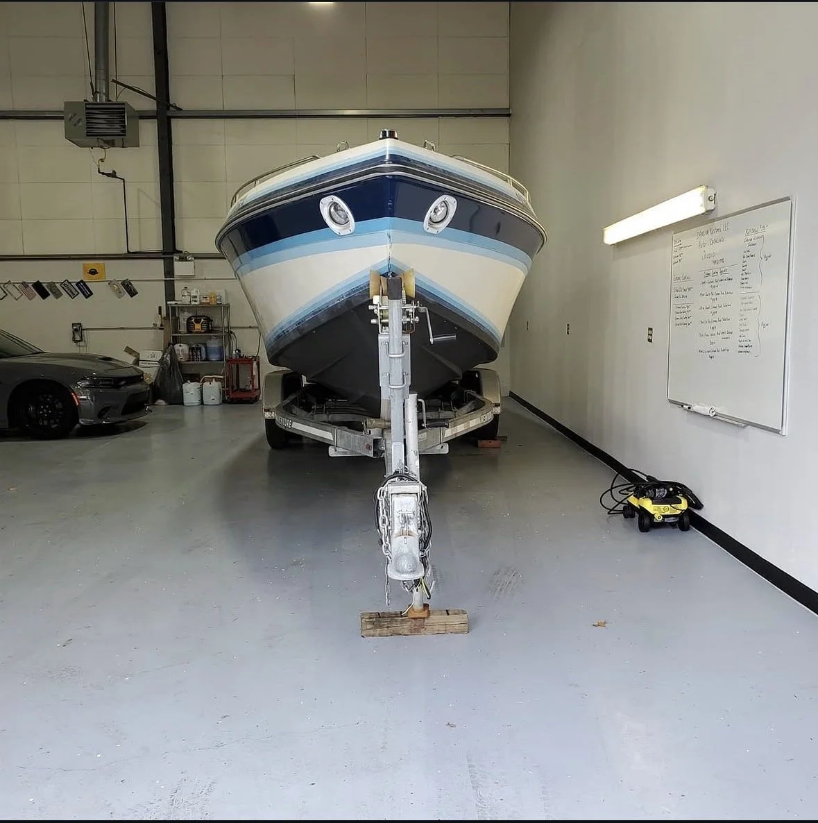 Boat on a trailer inside a garage with a whiteboard on the wall and a black car parked to the left.