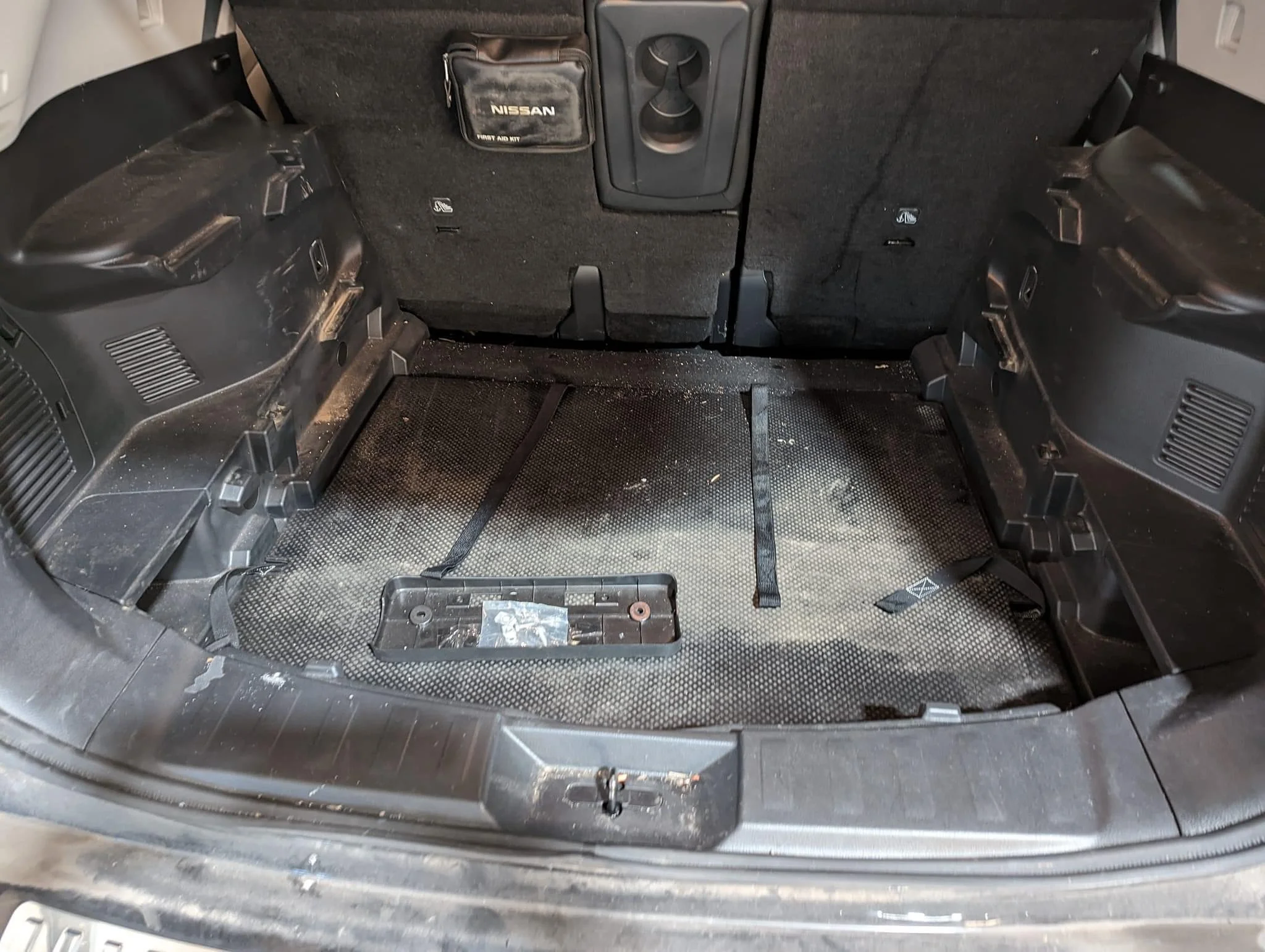 The back of a Nissan vehicle's interior with the rear seats folded down, showing a dirty and worn cargo area with straps and a plastic panel on the floor.