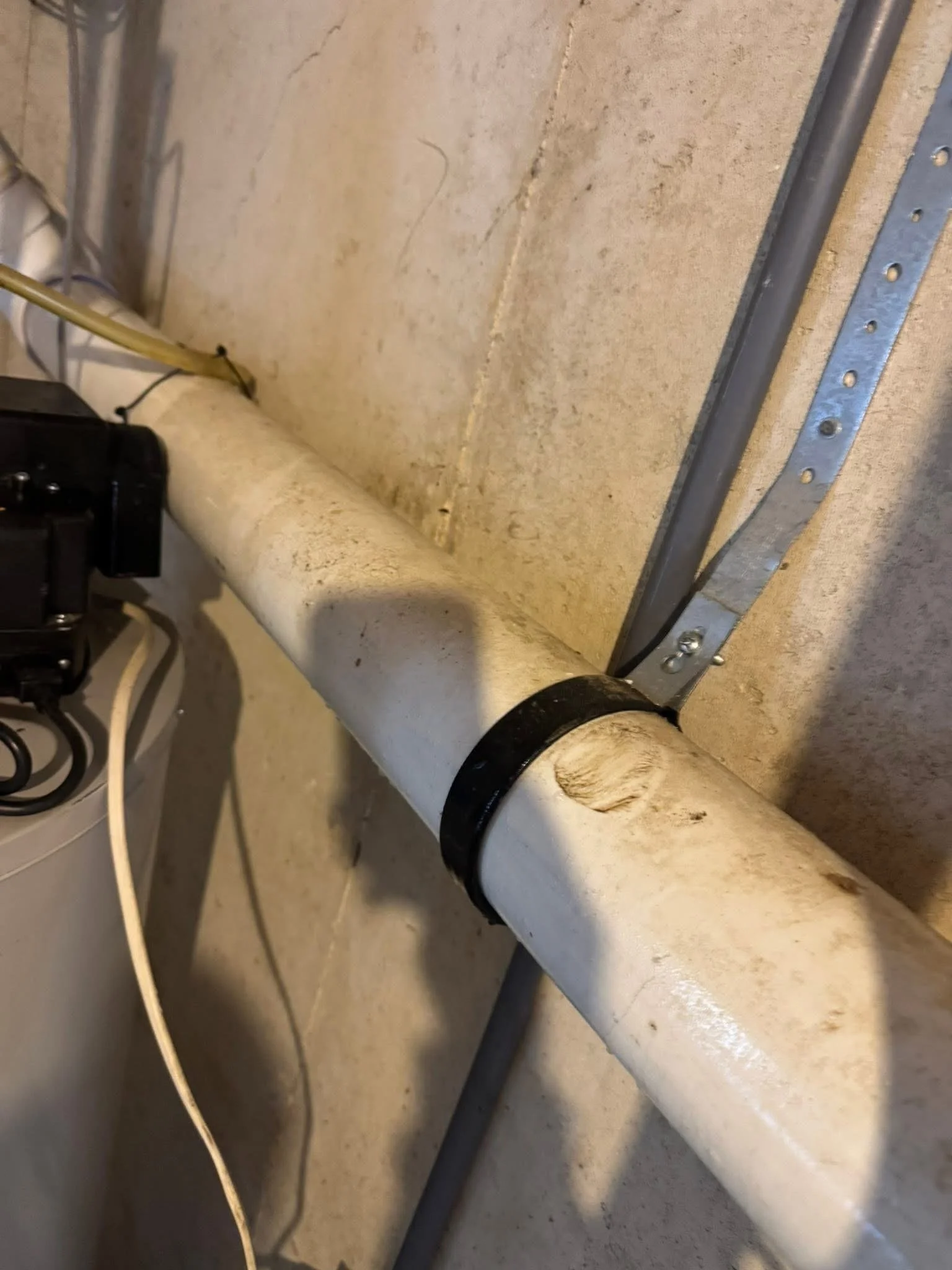 A white PVC pipe with a black coupler, mounted to a concrete wall with a metal bracket. Electrical wires and a grey conduit are also visible.