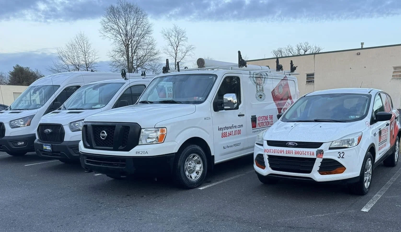 Parking lot with several vehicles, including a white fire protection service van, a white sedan, and other vans, with a building and leafless trees in the background.