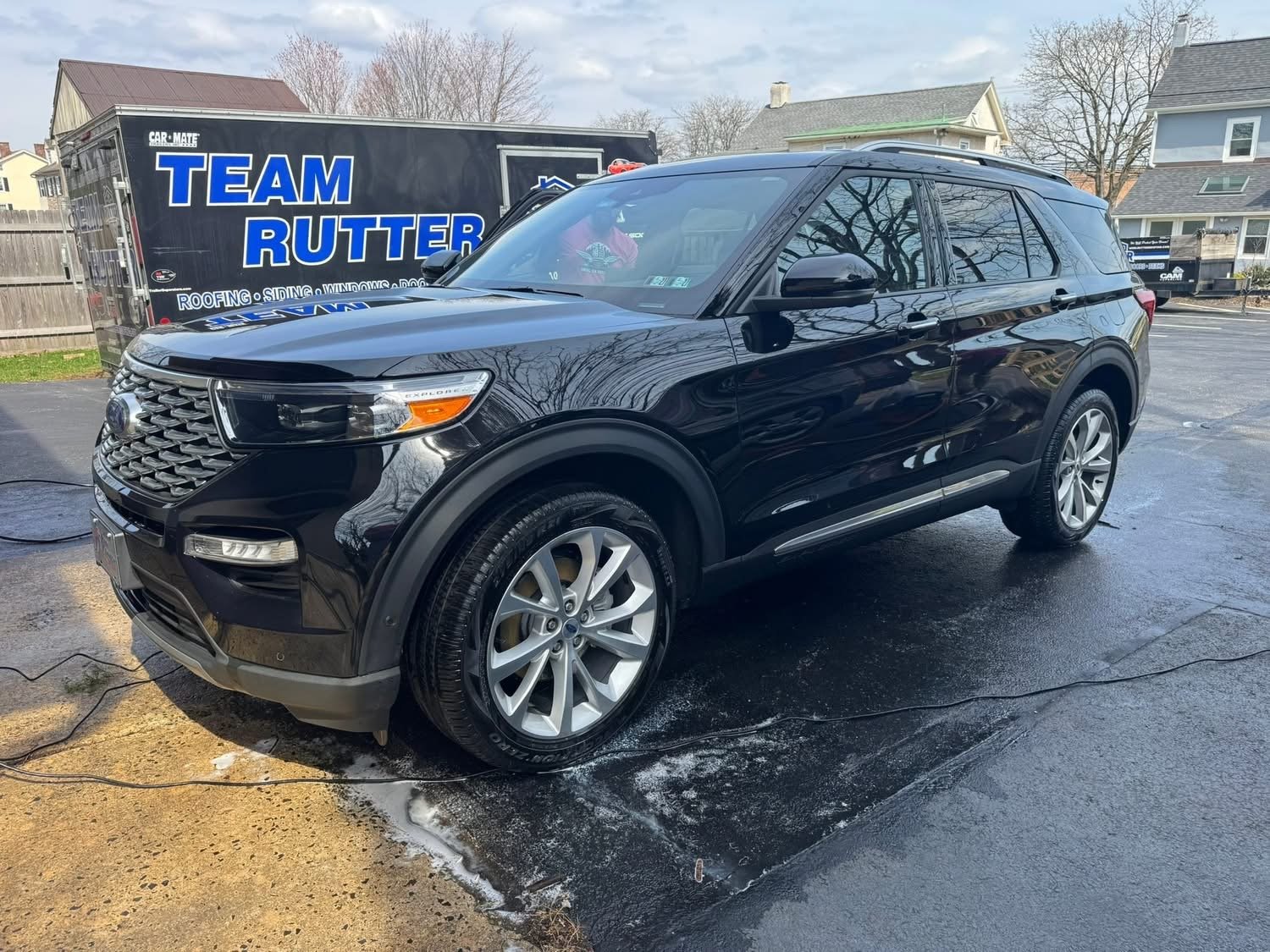Black SUV parked on a wet driveway with a gardening hose nearby, and a trailer with 'Team Rutter' branding in the background, residential houses and leafless trees also visible.