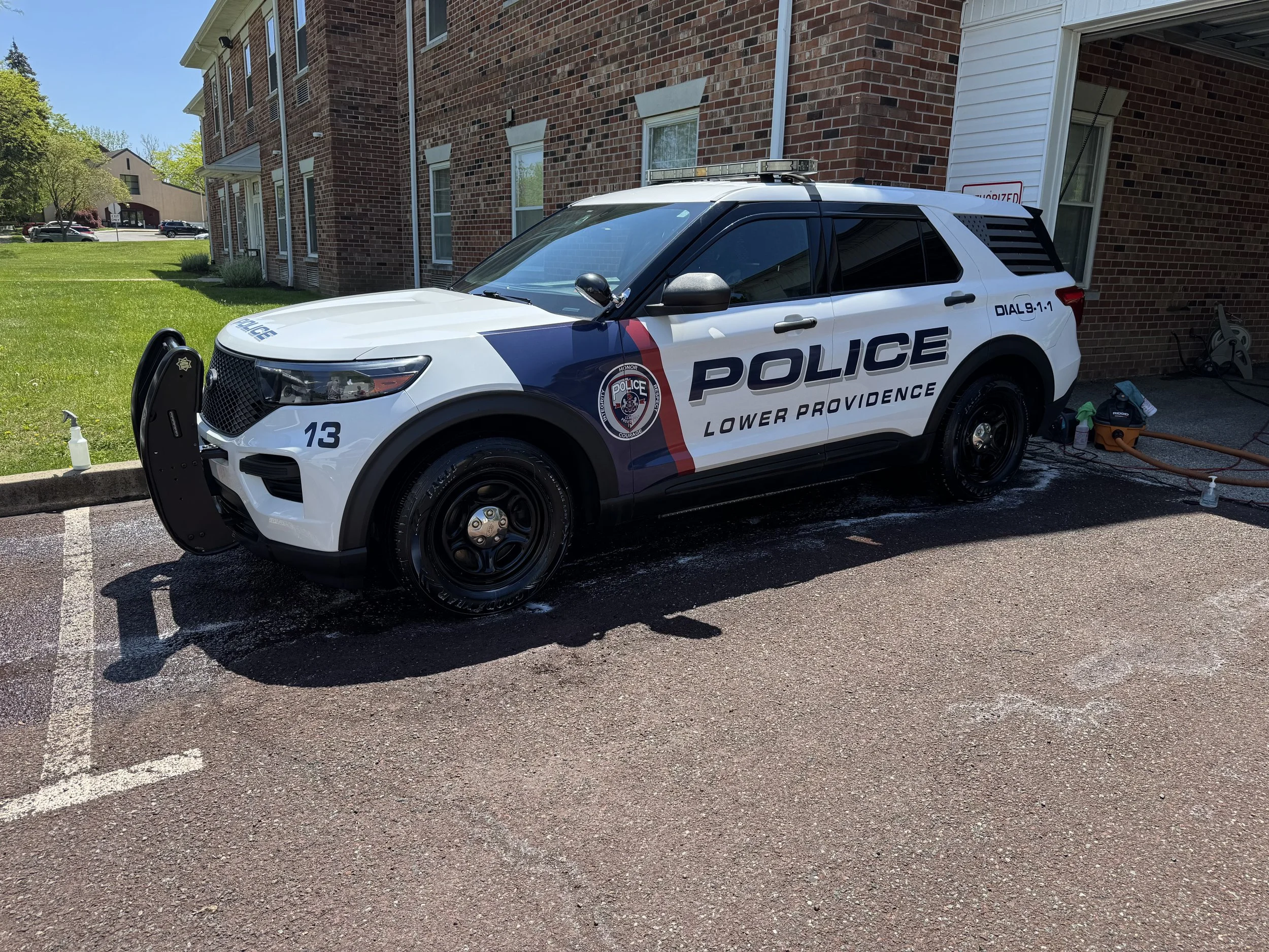 White police SUV parked outside a brick apartment building with a lawn. Equipment and cleaning supplies are visible on the ground near the vehicle.