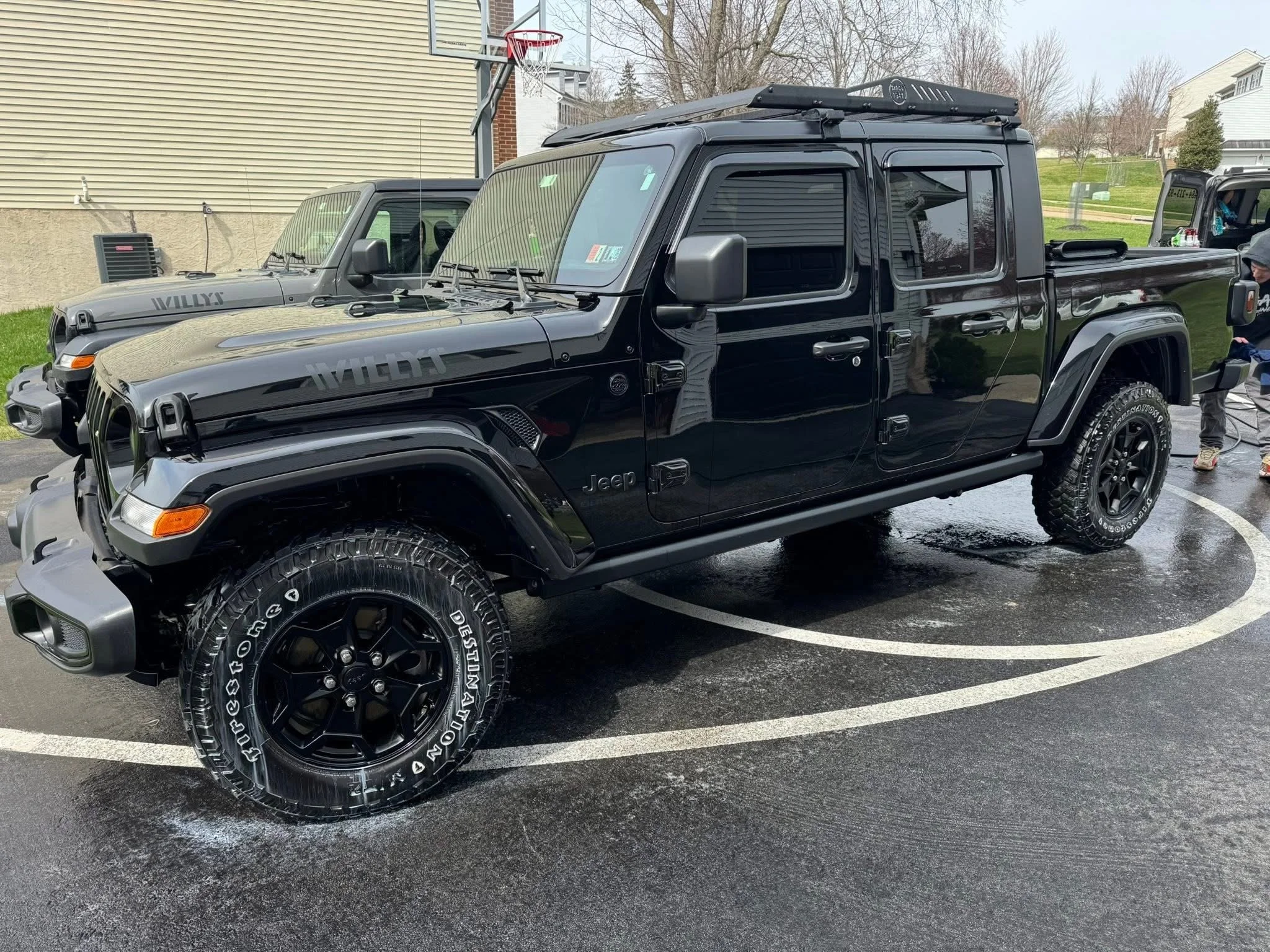 Black Jeep Gladiator pickup truck parked on wet asphalt, with a beige building and another vehicle in the background, and children playing nearby.