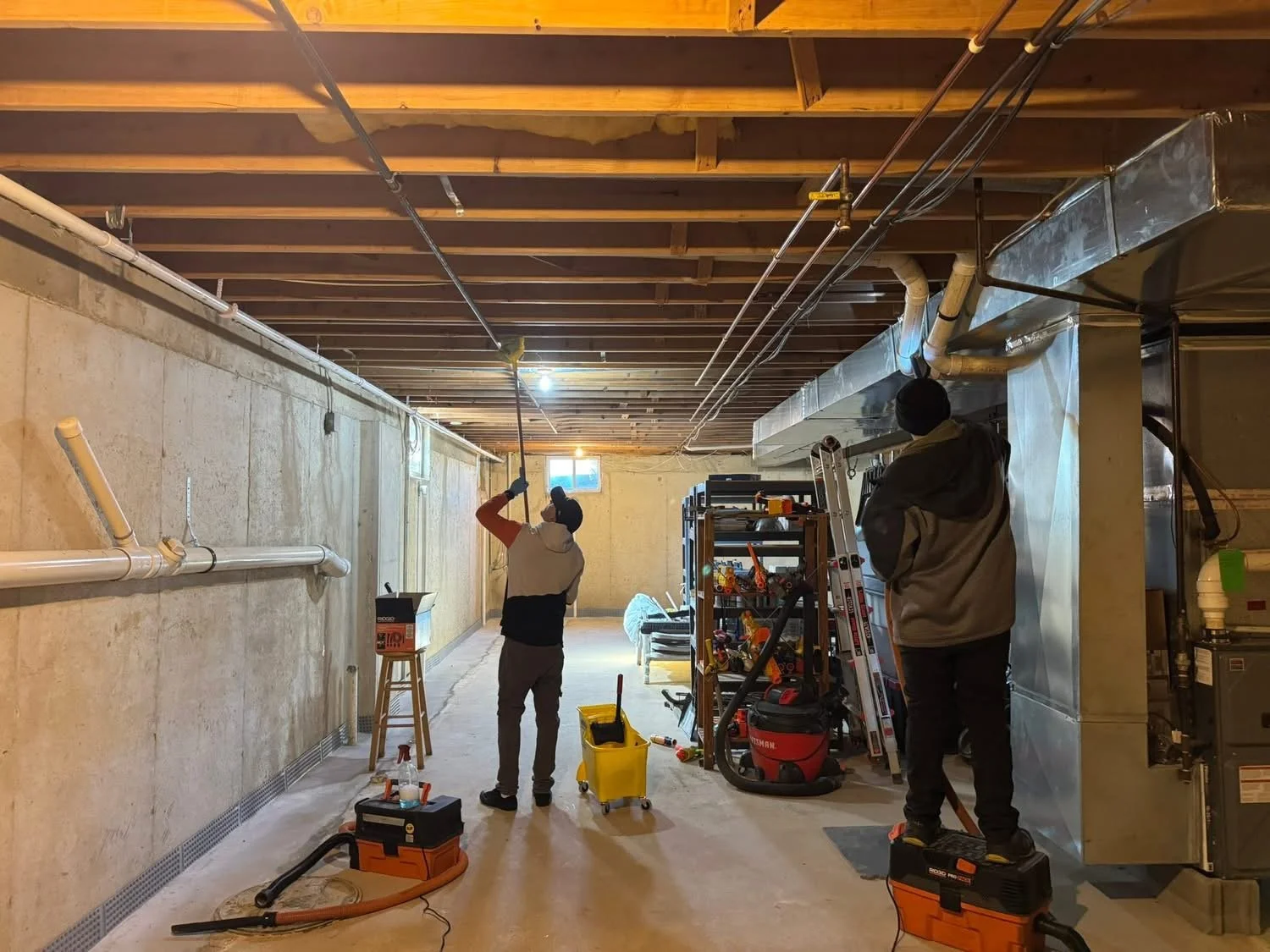 Two workers installing or repairing pipes in an unfinished basement with exposed ceiling beams and ductwork, using tools and equipment.