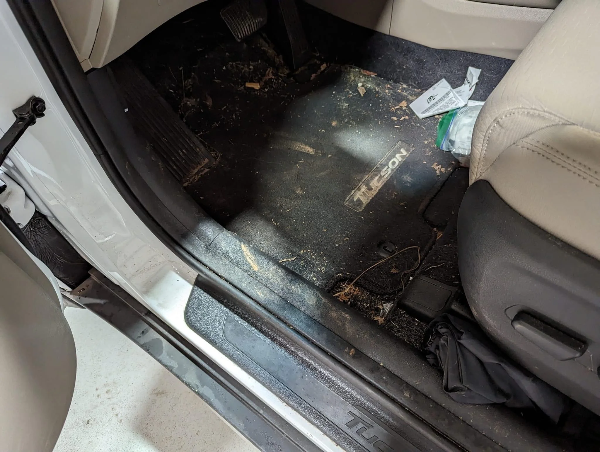 Dirty car floor mat inside a vehicle with debris and trash scattered around, including paper and a plastic bag.