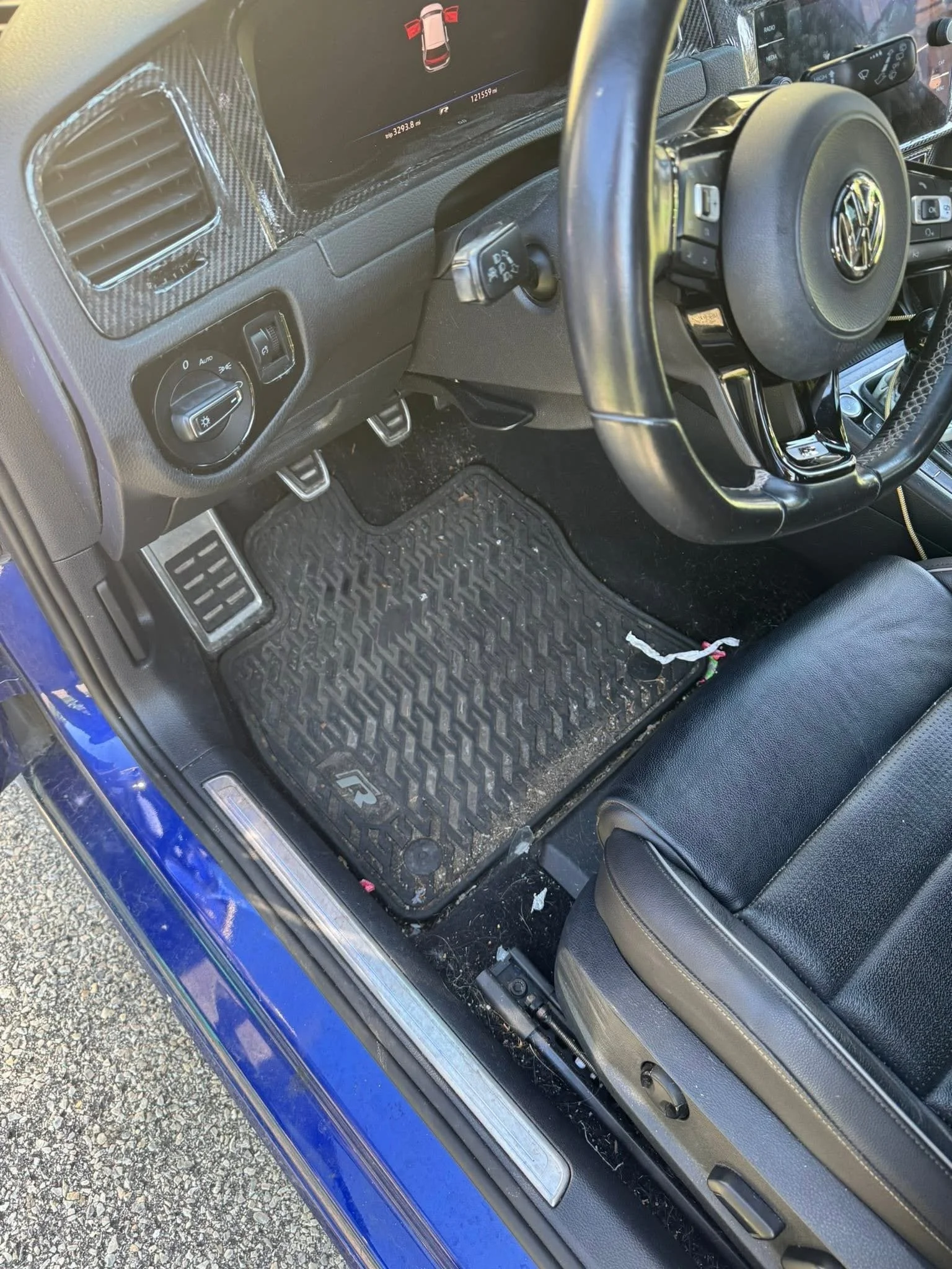 Inside a blue car, showing the driver's side floor area with a rubber floor mat, the gear shift, steering wheel, and dashboard with various controls.