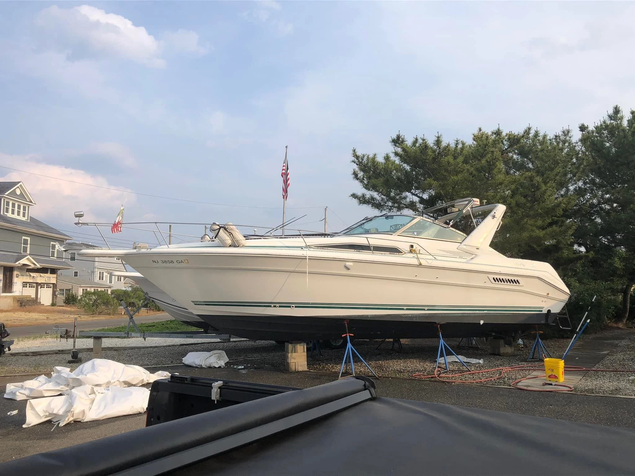 A white yacht is resting on a boat trailer in a backyard with houses and green trees. An American flag is flying in the background, and there are some white coverings and equipment on the ground.
