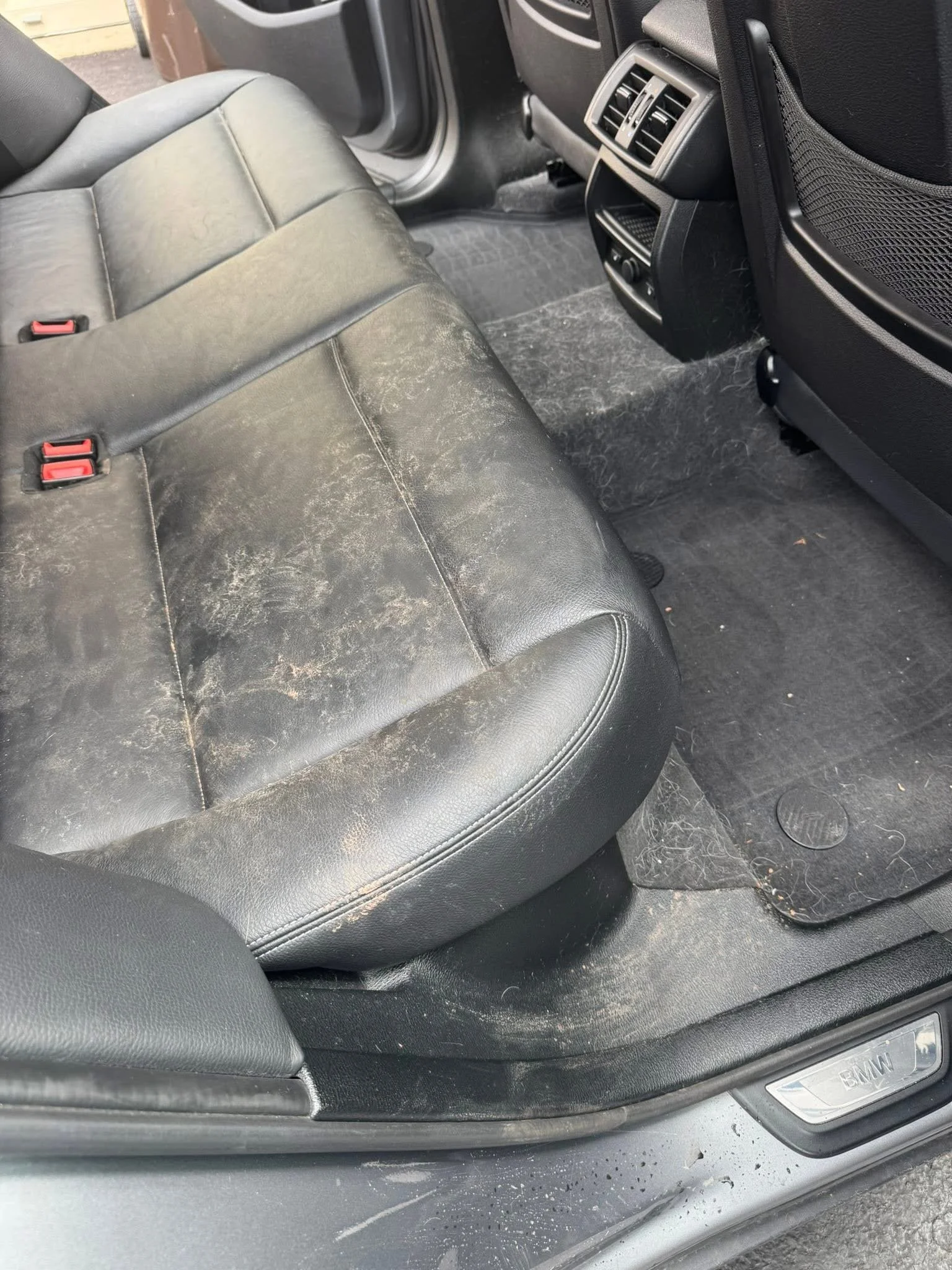 The rear passenger seat and floor of a black leather car interior, showing dirt and debris on the seat and floor mats.