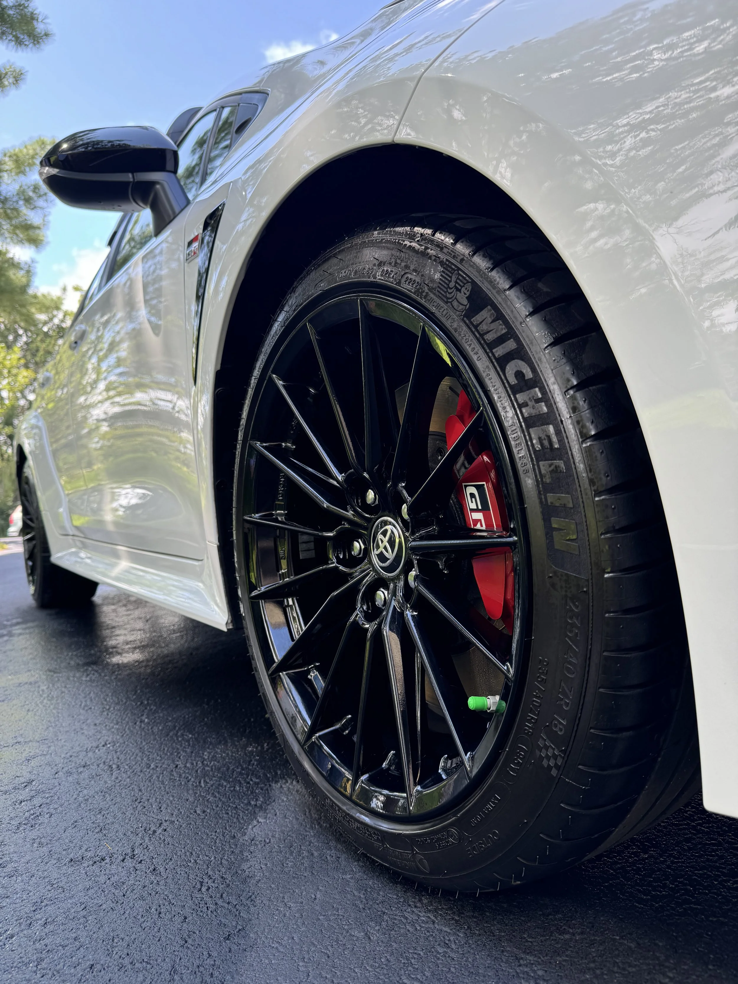 Close-up of a white sports car wheel with black rim and red brake caliper, parked on a wet asphalt surface in sunlight.