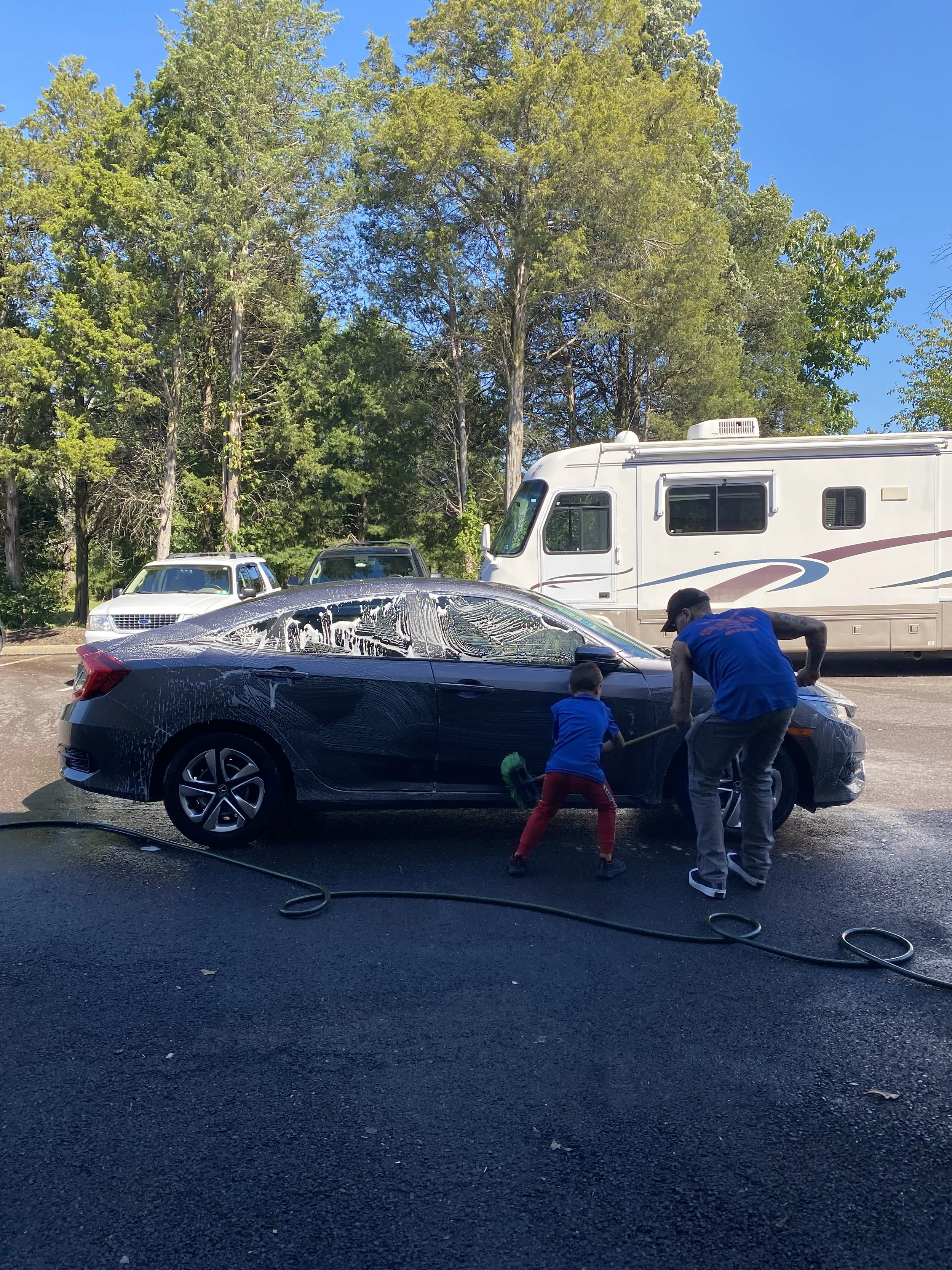 A man and a young boy washing a black car with soap and water, outdoors on a sunny day. There are trees and parked vehicles, including a large RV, in the background.