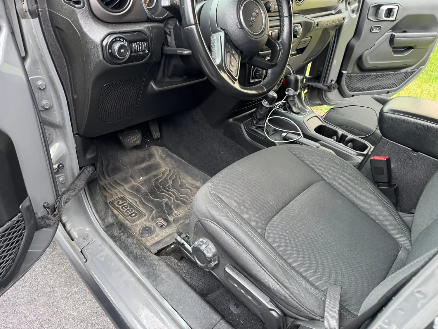 Interior of a Jeep vehicle showing the driver's seat, steering wheel, dashboard, and center console with gear shift, cup holders, and electronic accessories.