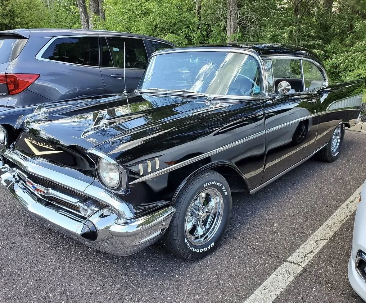 A black vintage Chevrolet car parked in a lot next to a modern grey SUV with trees in the background.