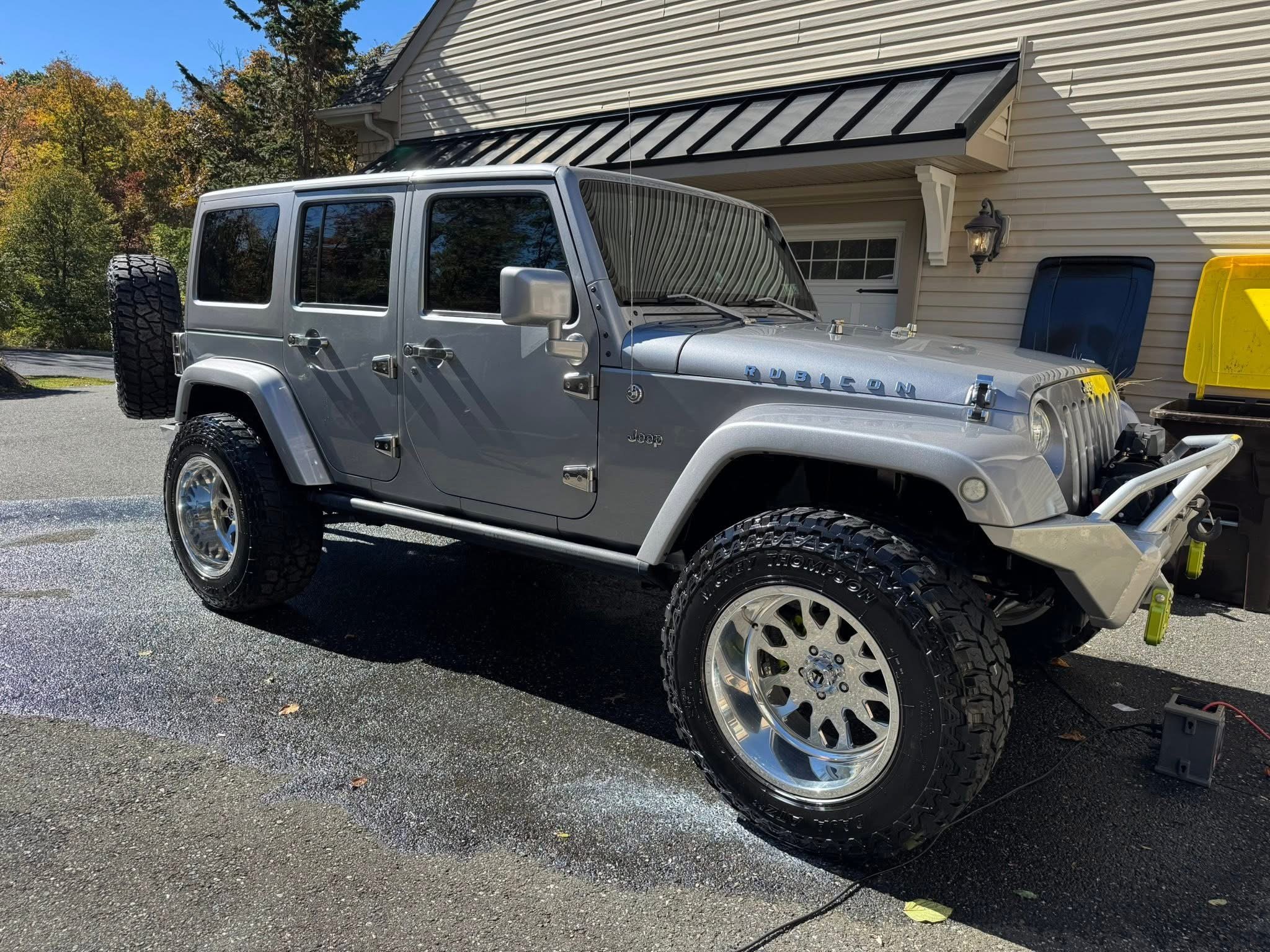 A silver Jeep Rubicon parked on a driveway, with a spare tire mounted on the rear, large off-road tires, and a white stripe on the hood. The background includes a house with a garage, a yellow trash bin, and trees with fall foliage.