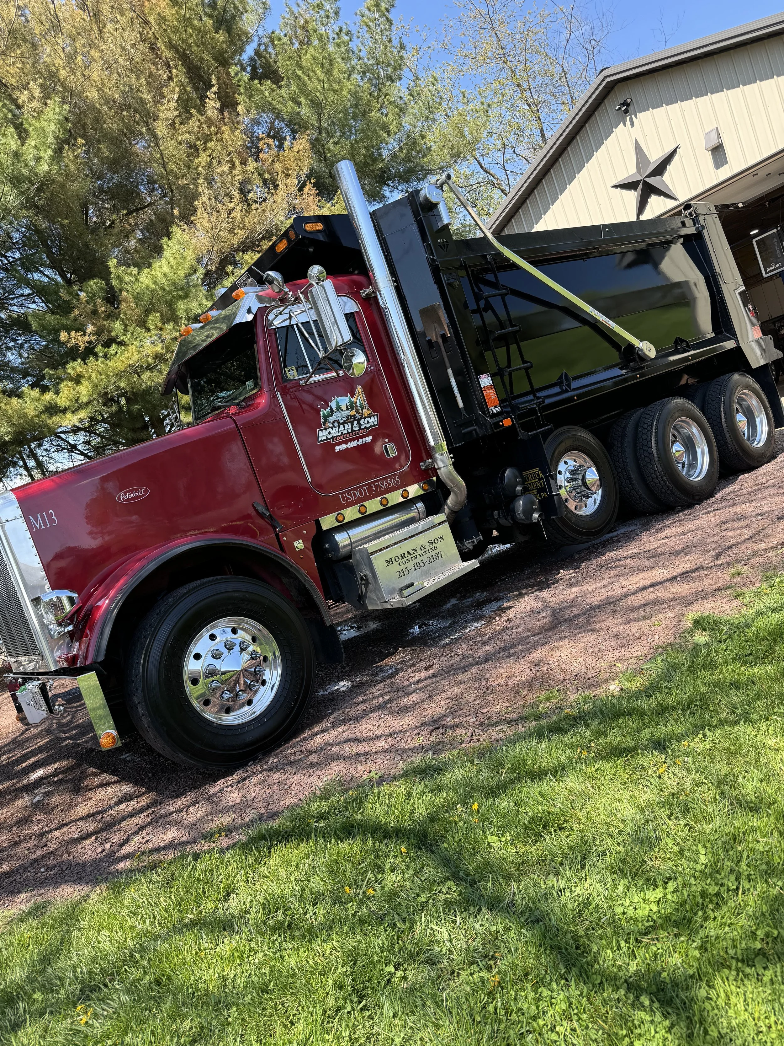 Red and black dump truck parked on a gravel surface next to green grass. A building with a metal roof and star decoration is in the background, and trees are behind the truck, with a clear blue sky above.