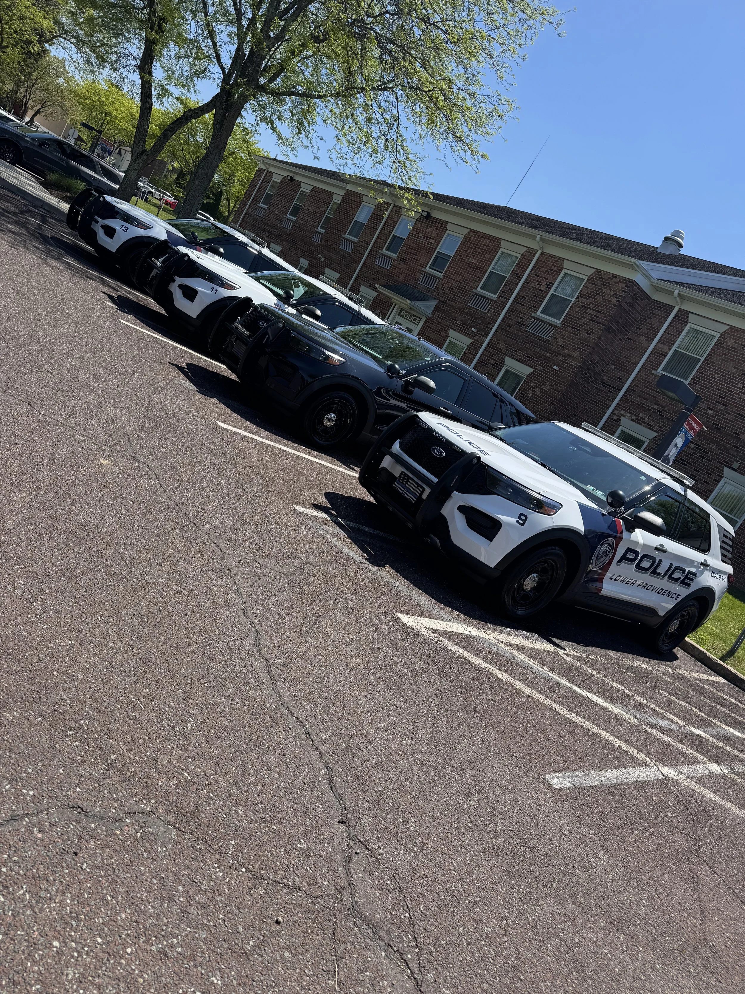 Four police cars parked in a parking lot in front of a brick building with multiple windows and a police sign. Trees with green leaves are visible in the surroundings, under a clear blue sky.