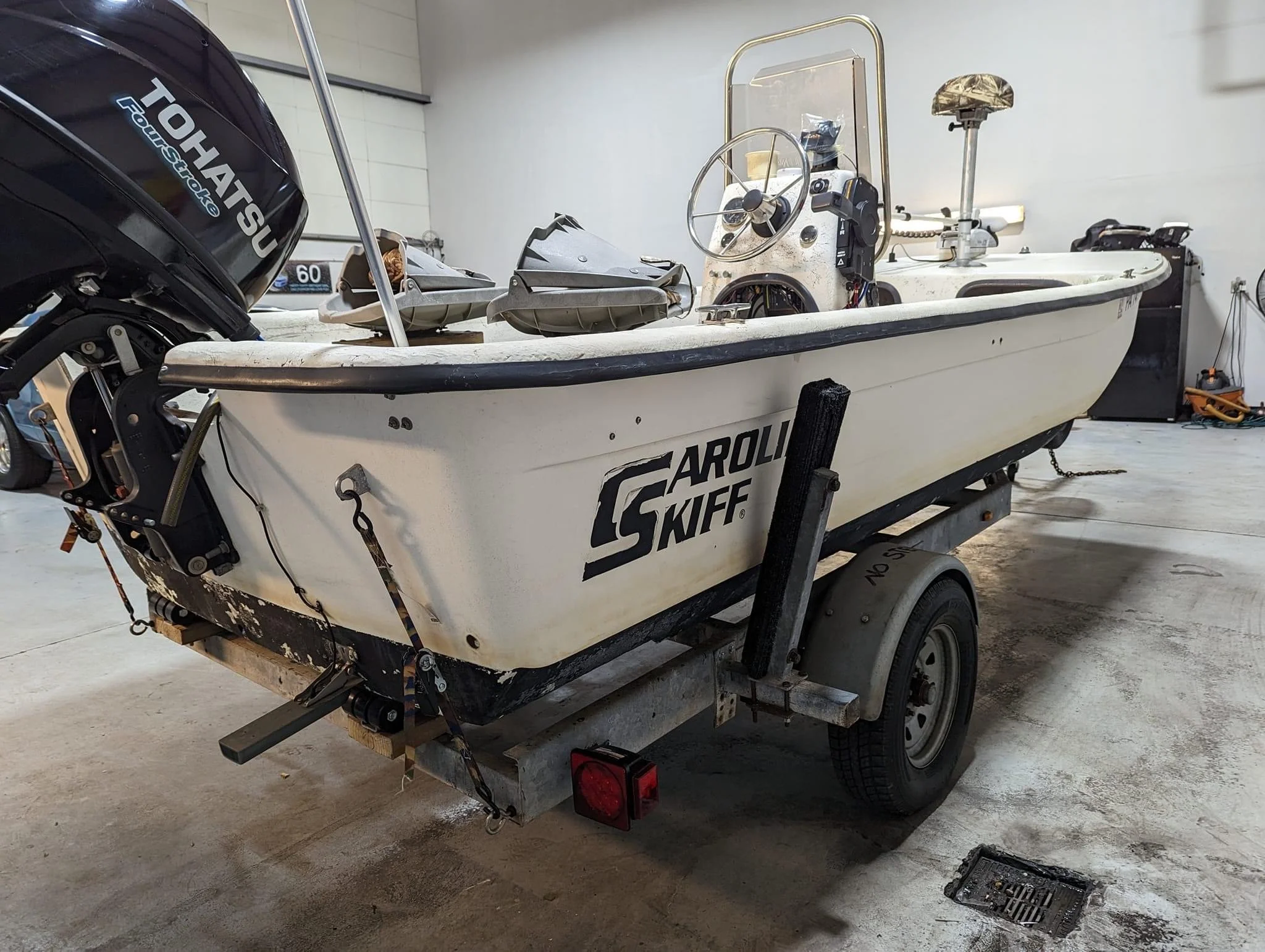 A small white boat with a black outboard motor attached to the back, resting on a trailer in an indoor space. The boat has a steering wheel and some equipment on the console. The space appears to be a garage or warehouse.