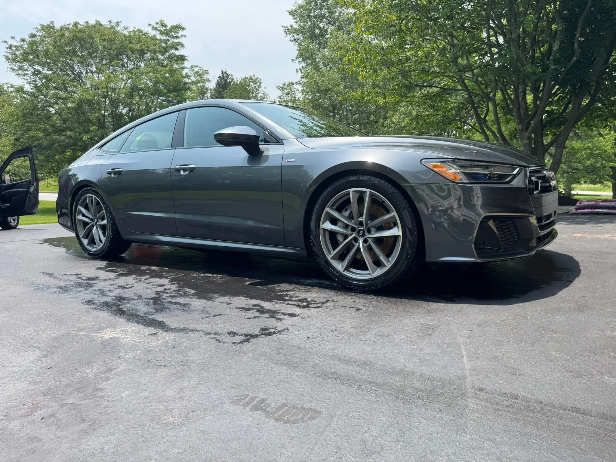 A sleek, silver sedan car with tinted windows parked on a wet asphalt surface, with a open passenger door and a puddle nearby, surrounded by green trees and a cloudy sky.