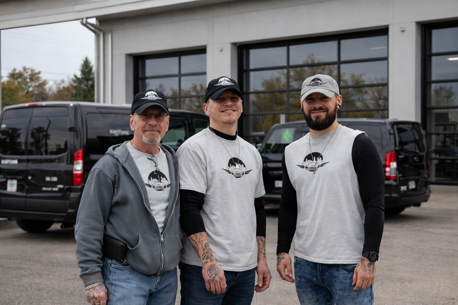 Three men standing outdoors in front of a building with black vans in the background. All wearing grey and black clothing with matching logos on their shirts and caps.