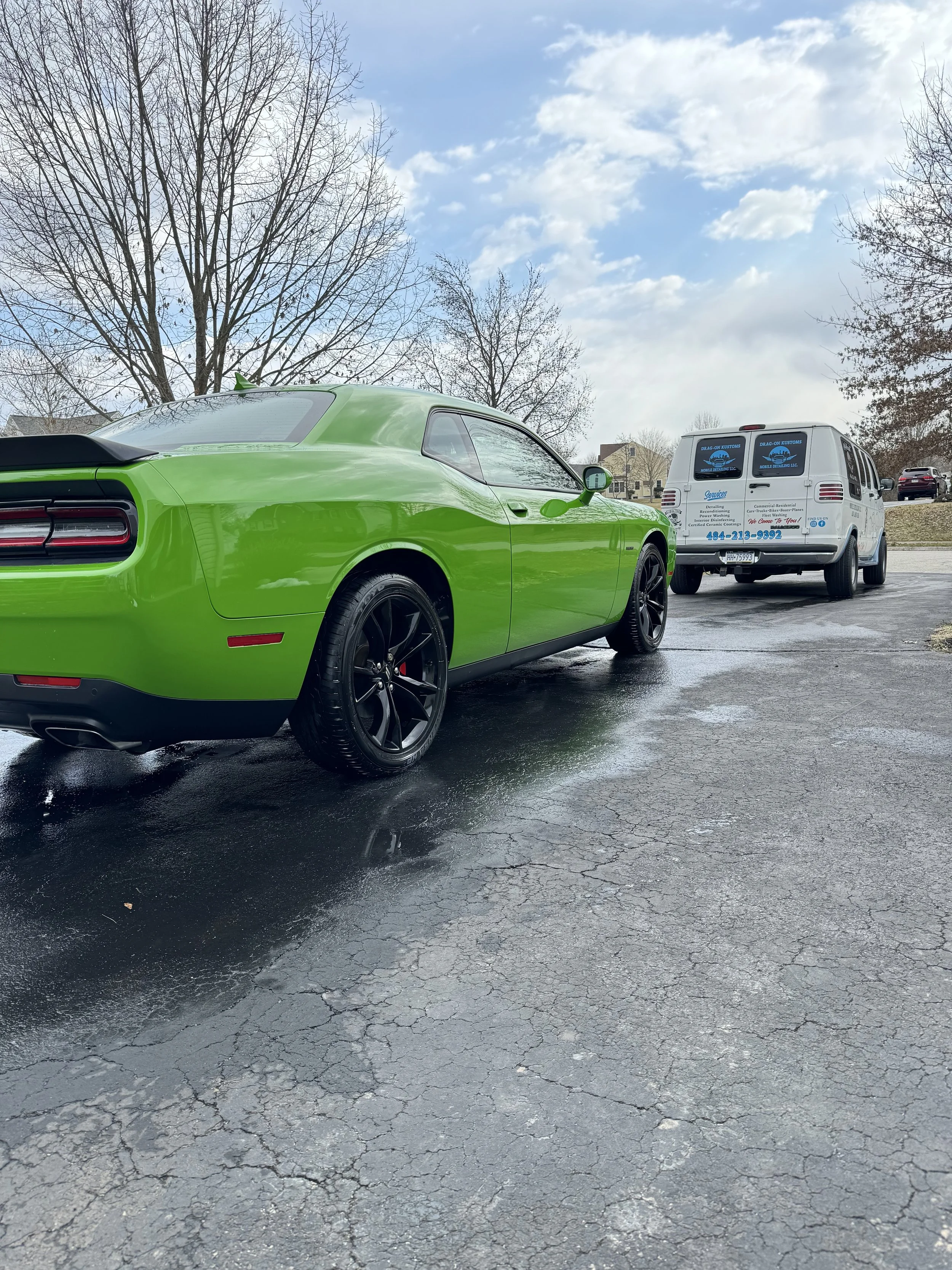 A bright green modern sports car with dark wheels parked on a wet asphalt surface, with a white service van in the background and leafless trees under a partly cloudy sky.