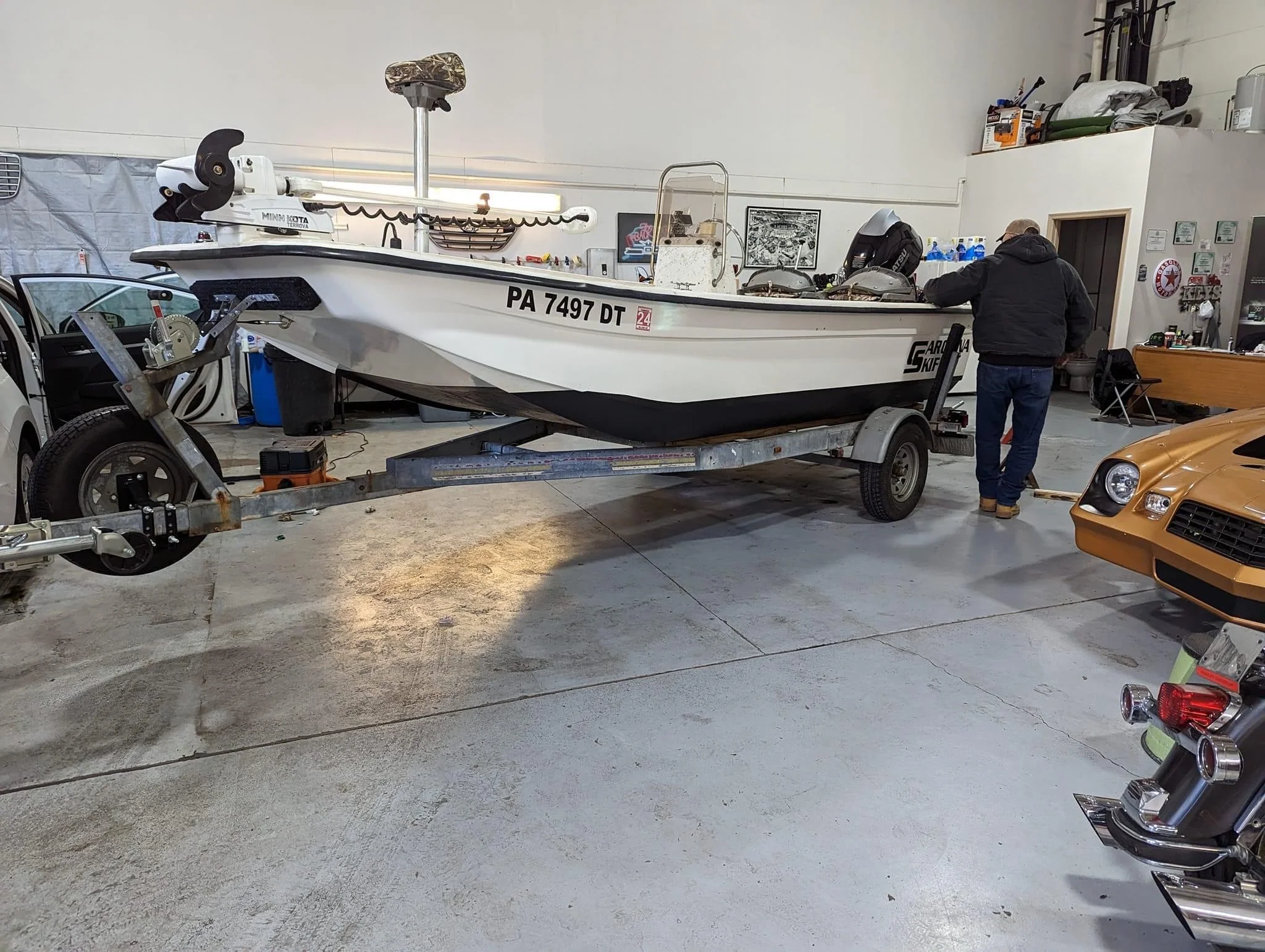 An indoor garage with a large white boat on a trailer, a man working on the boat, and various tools and items around.