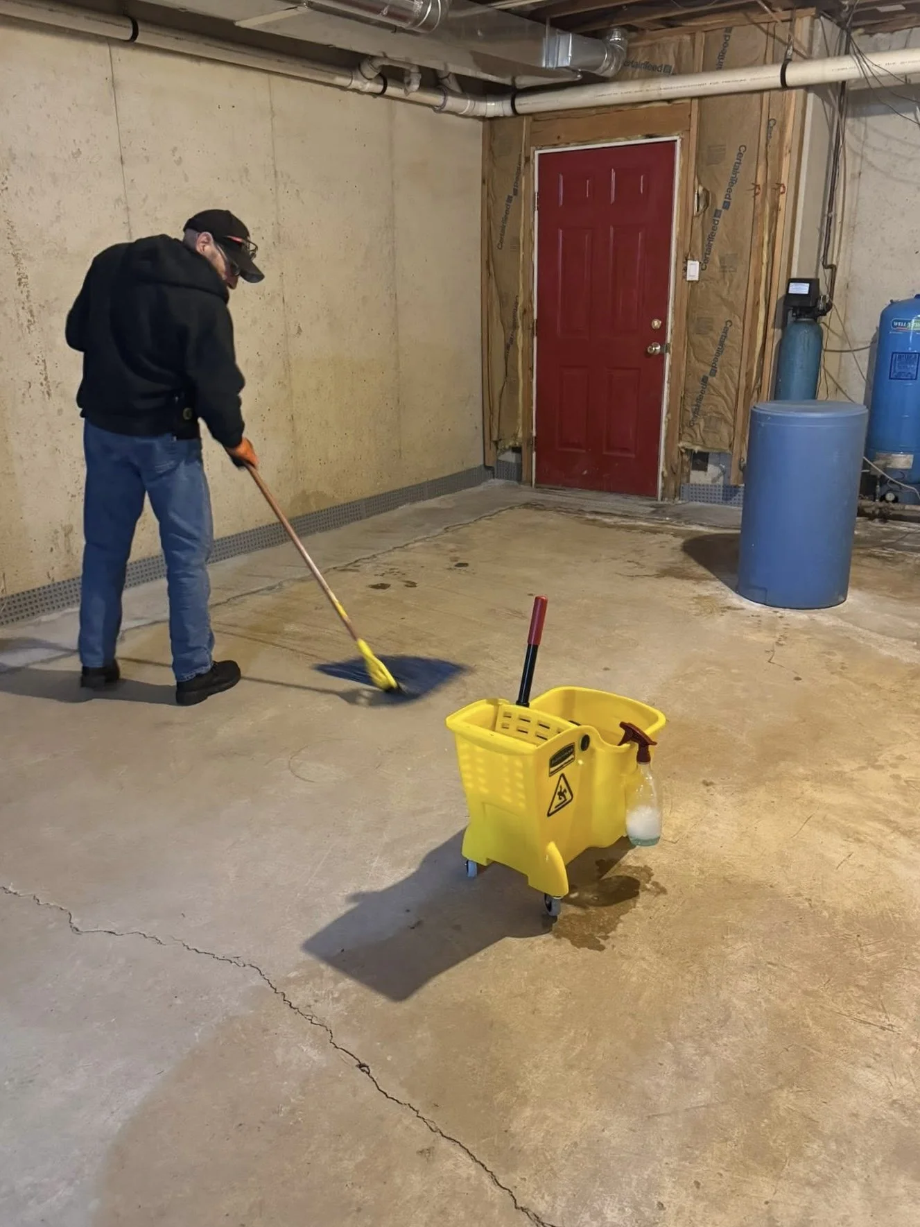 Person mopping concrete floor in a garage with cleaning supplies and equipment visible in the background.
