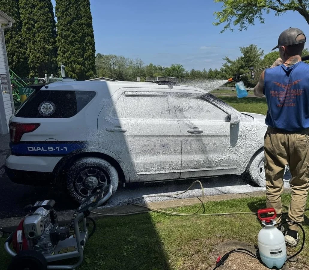 Person pressure washing a white police car with foam, outside on a sunny day.