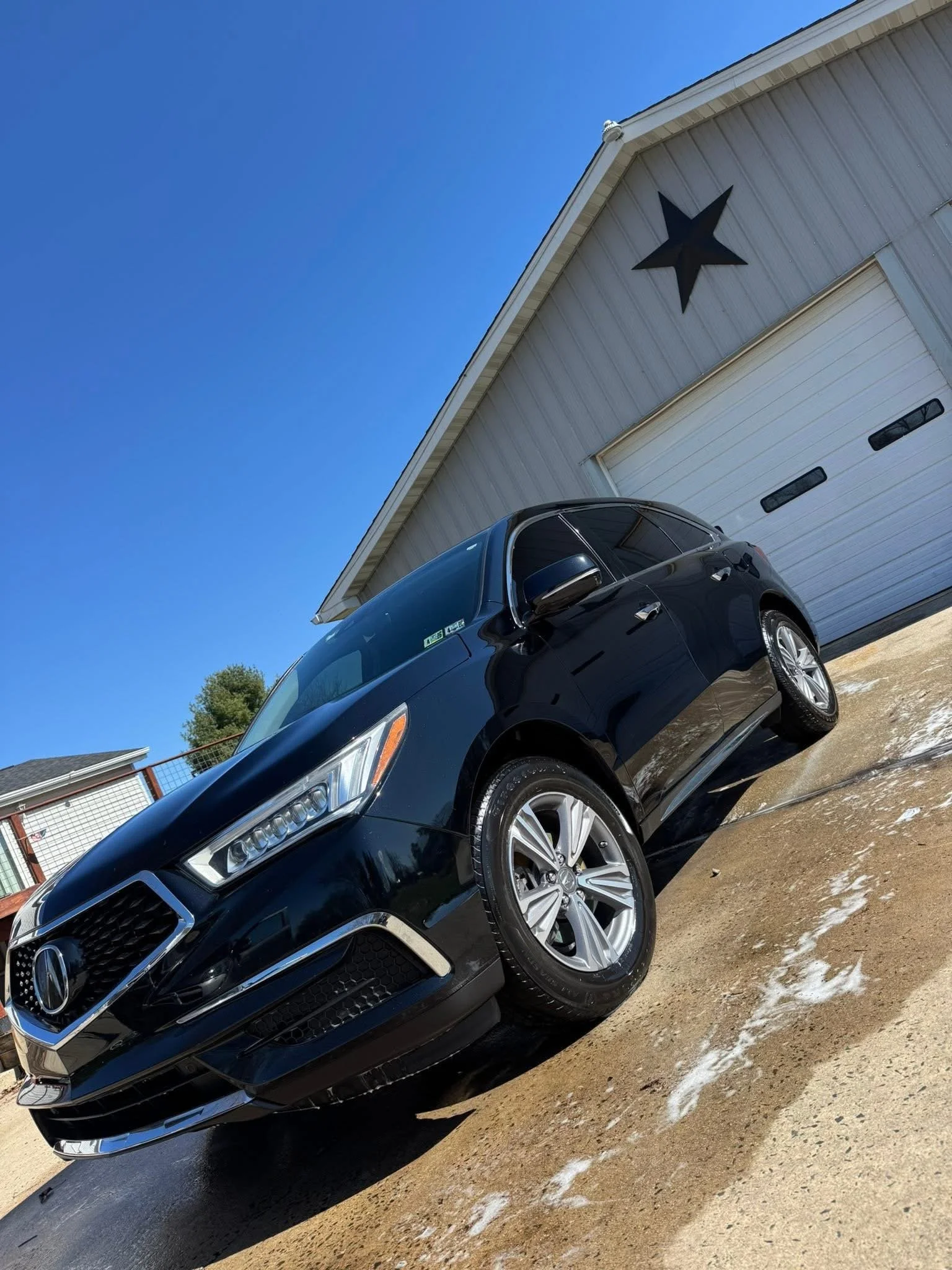 Black Acura SUV parked in front of a white garage door with a black star decoration on the house exterior, under a clear blue sky.