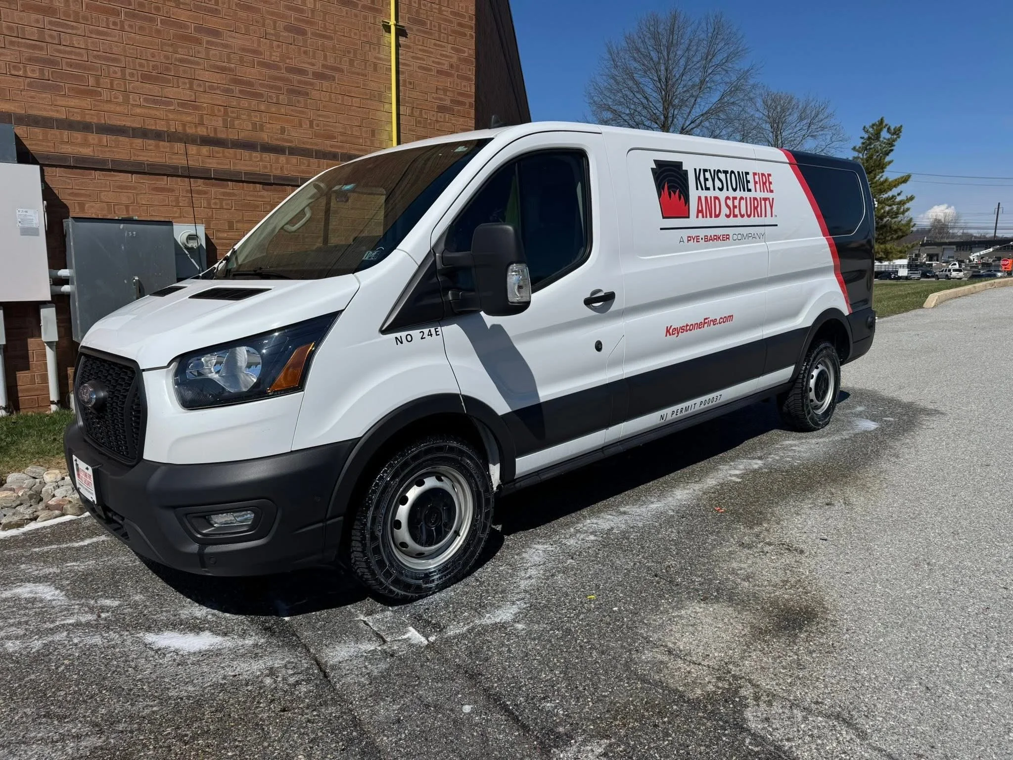 White service van with 'Keystone Fire and Security' logo parked near a brick building under a clear blue sky. Trees and a parking lot are visible in the background.