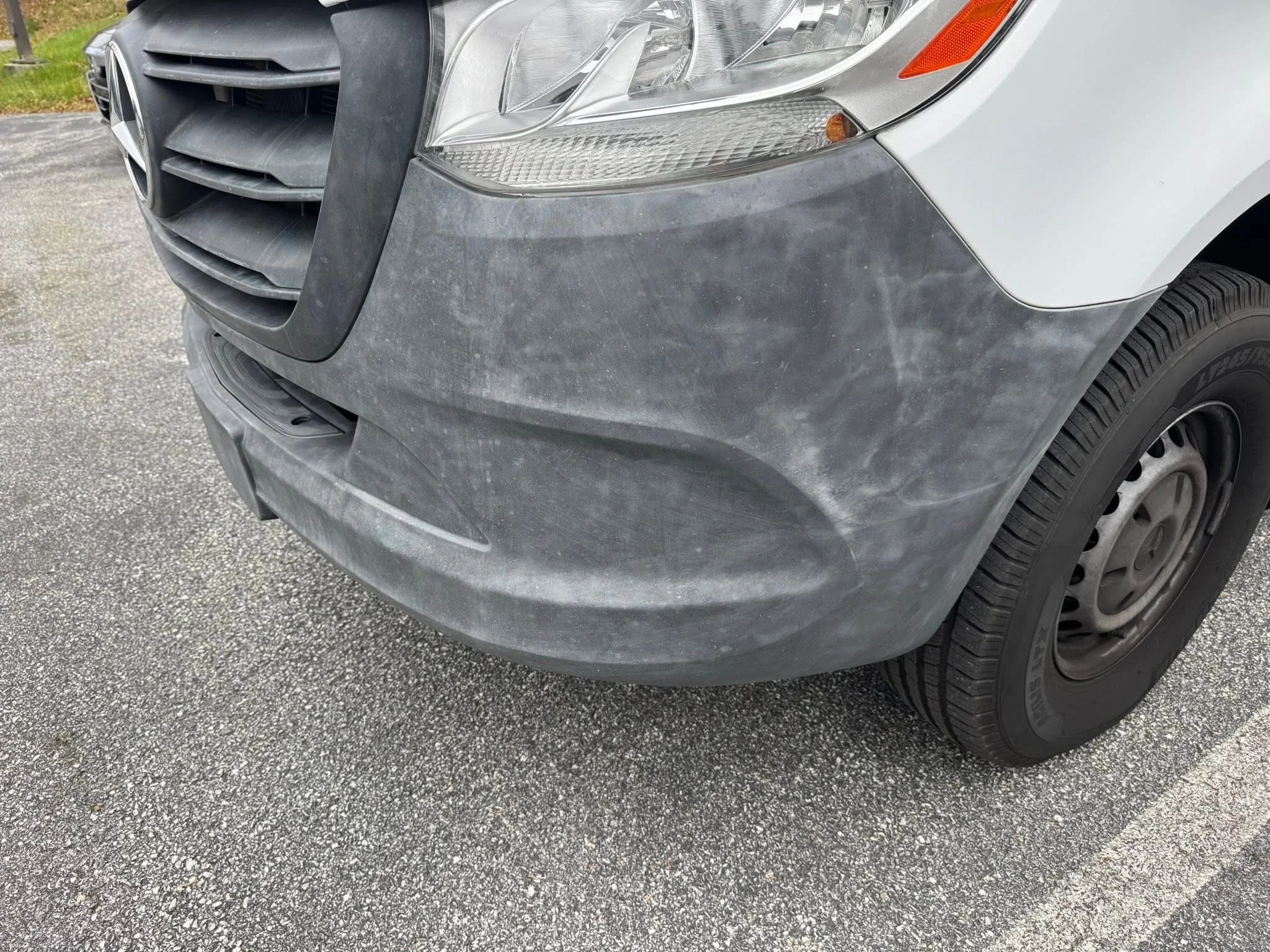 Close-up of the front right side of a gray van with visible dirt and scratches on the bumper and fender, parked on a paved surface.