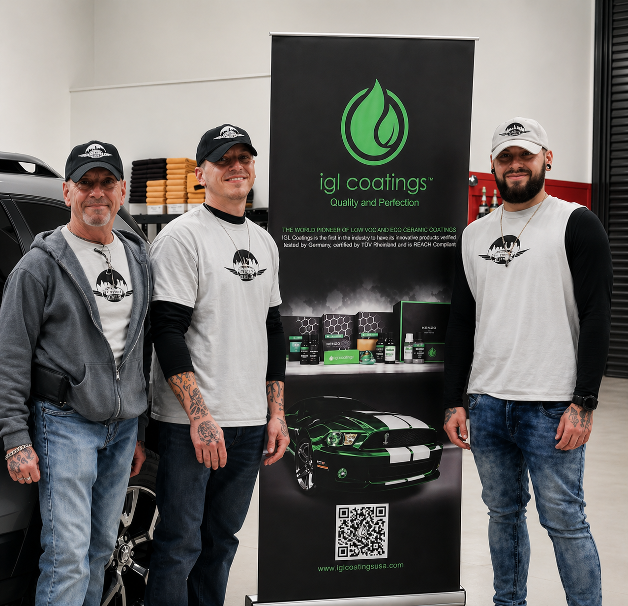 Three men standing beside a promotional banner for IGL Coatings, a company specializing in low VOC and eco-friendly ceramic coatings. The men are smiling, wearing hats and casual clothing with the company's logo. The banner features a green logo, product images, a QR code, and a black and green sports car illustration.