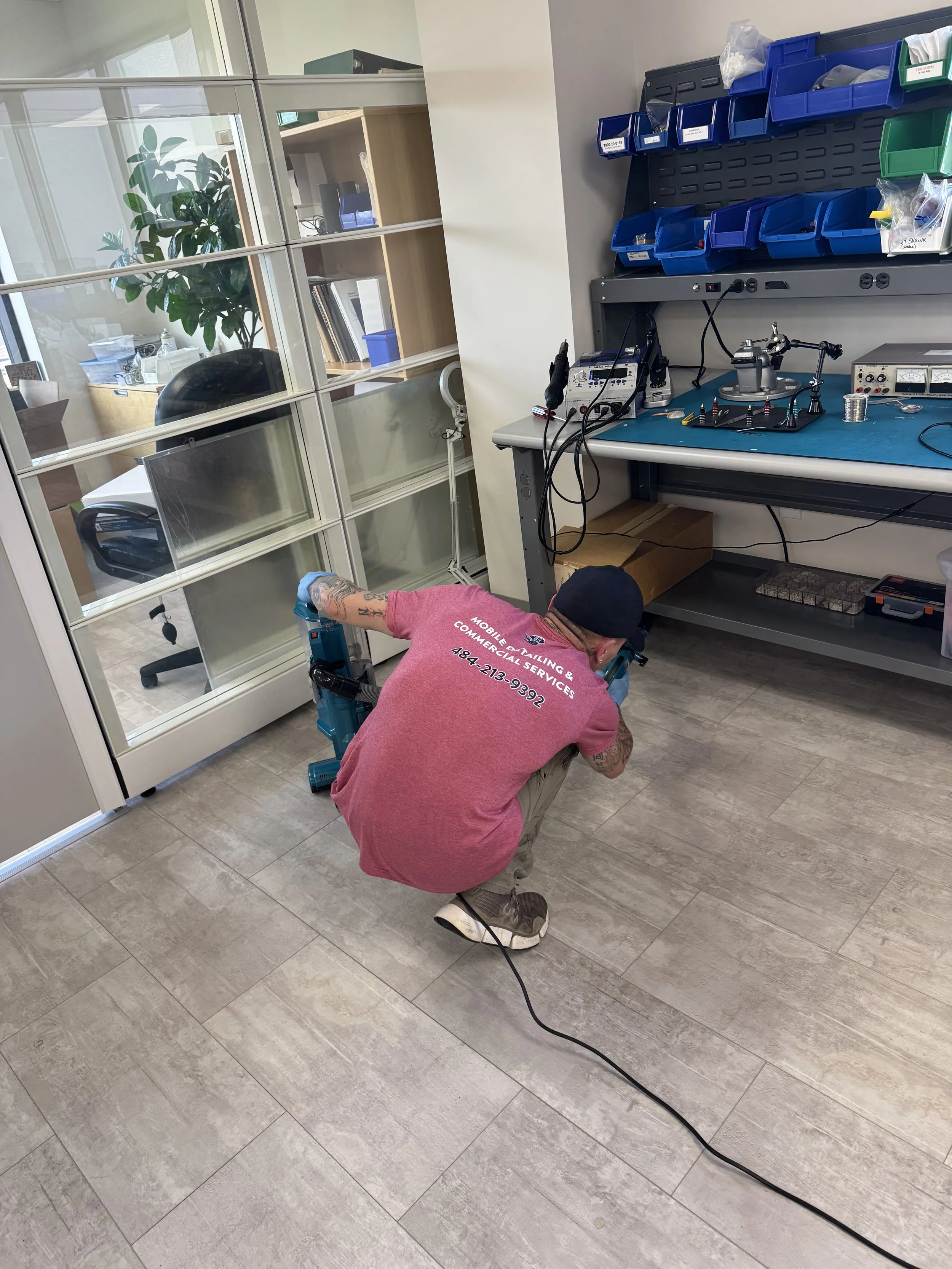A technician with tattoos, wearing a red shirt with service information on the back, is crouching on the floor working on a power drill in an office or workshop. The workspace includes a glass-walled office, a workbench with various tools, electronic