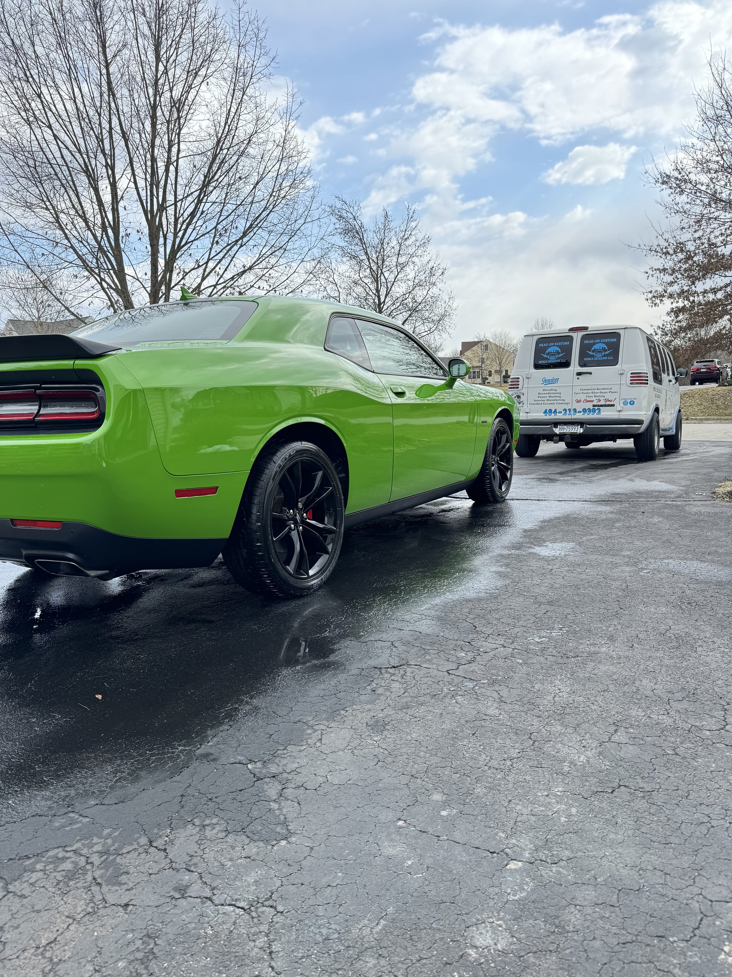 A bright green sports car parked on a cracked pavement with a white service van in the background and leafless trees under a partly cloudy sky.