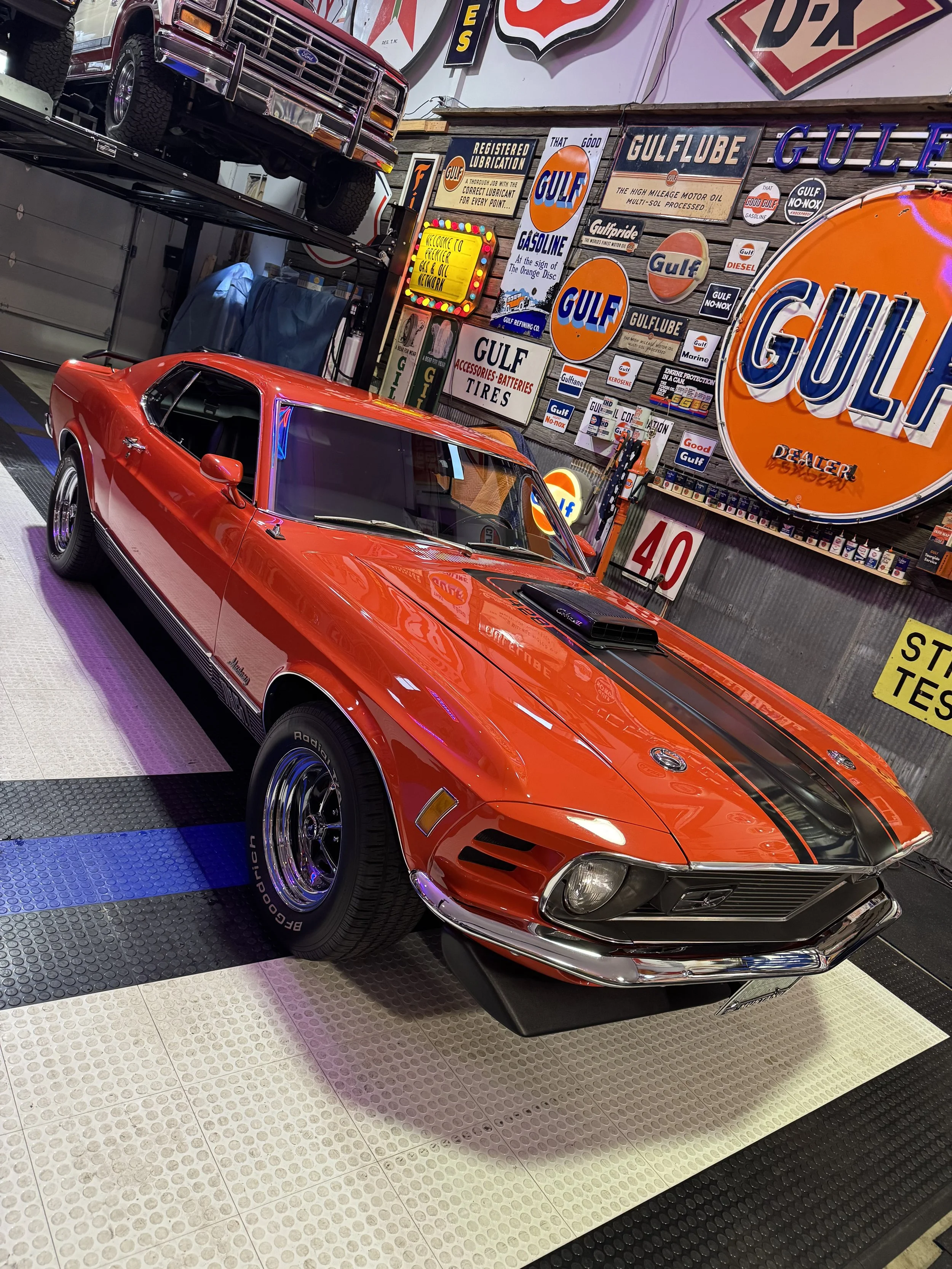 A red vintage Ford Mustang Shelby car with black racing stripes, displayed indoors with vintage Gulf oil signs on the wall behind it.