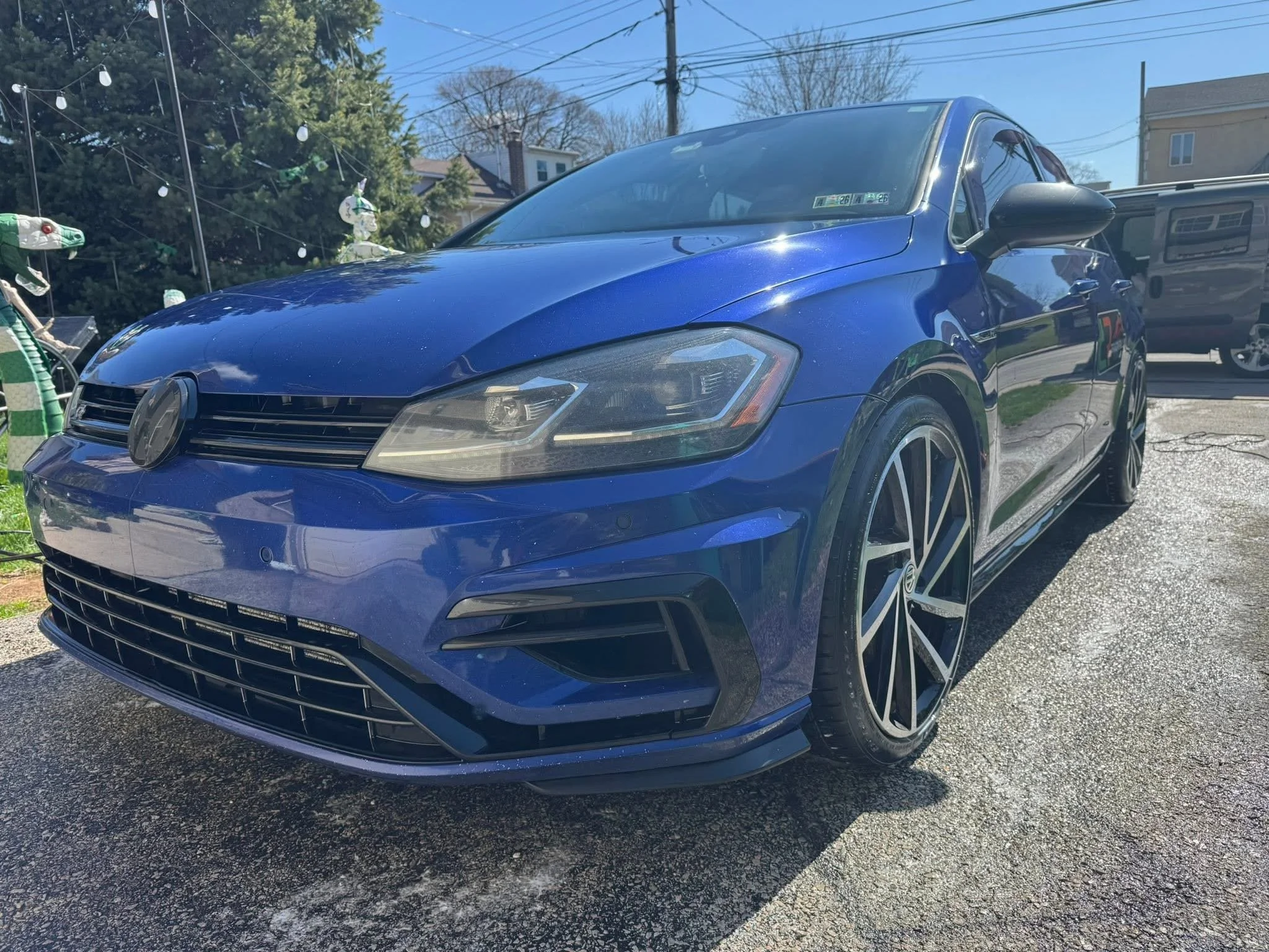 Blue Volkswagen sedan parked on a driveway with a background of trees, house, and other vehicles, under a clear blue sky.