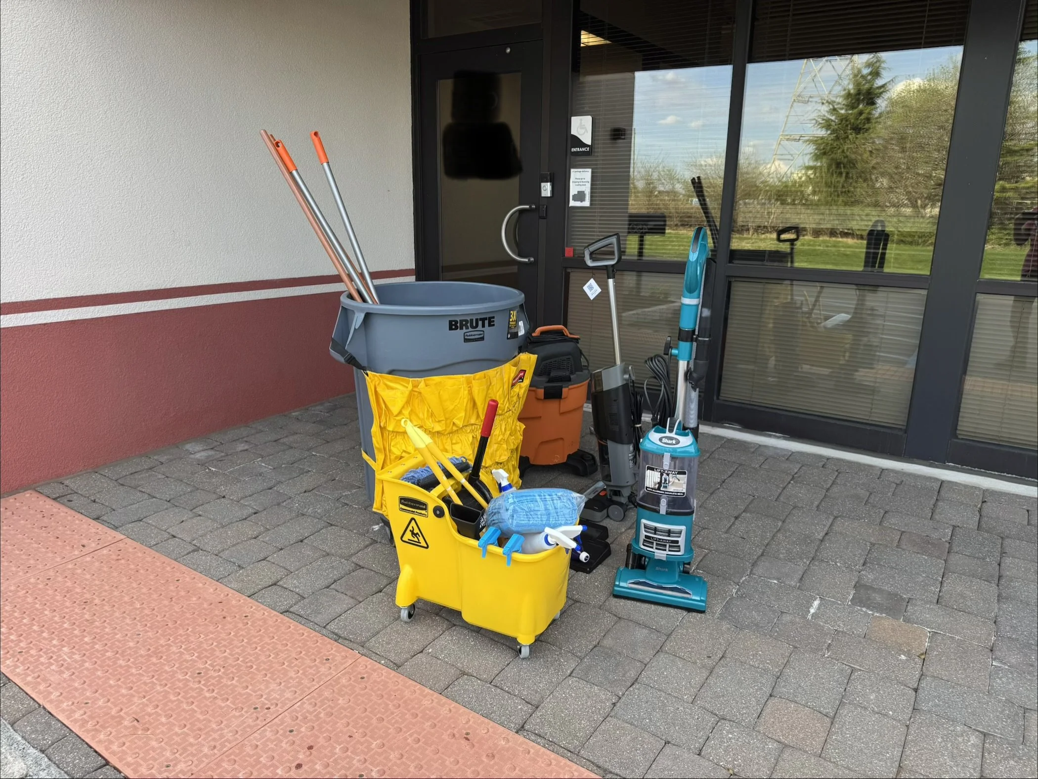 Cleaning supplies including mop, bucket, vacuum, and cleaning tools outside a building entrance.
