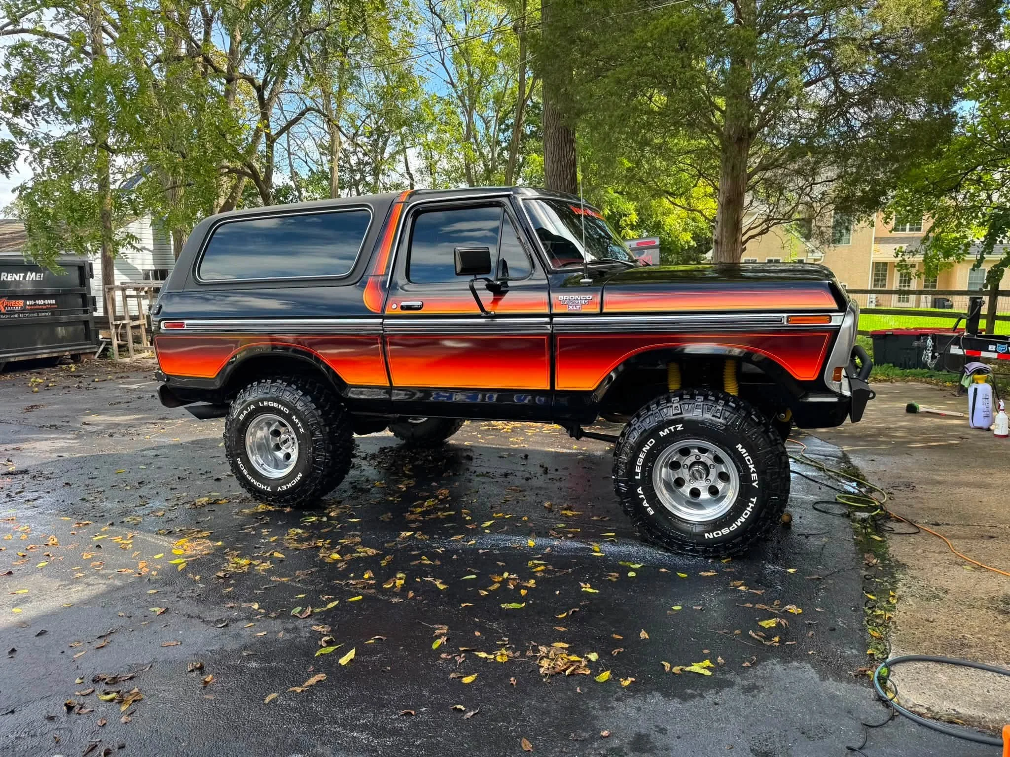 A black off-road vehicle with orange and red flame decals on the sides, sitting outdoors on a driveway wet with water, surrounded by trees and residential buildings.
