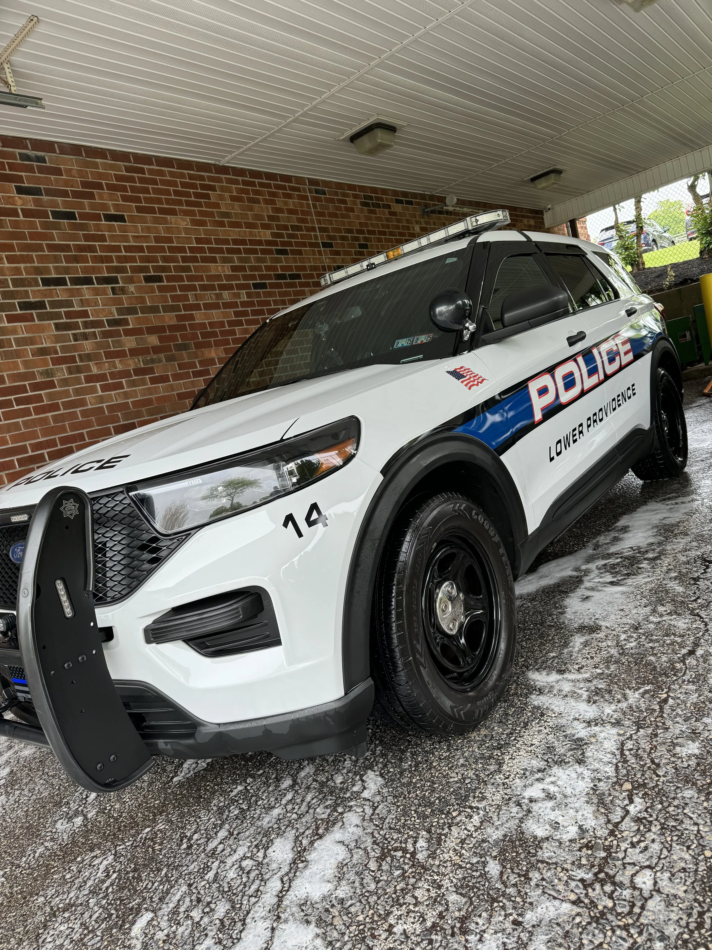 A police car parked in a covered area with a brick wall behind it and other vehicles visible outside. The police car is white with black and blue markings, and has a light bar on top.