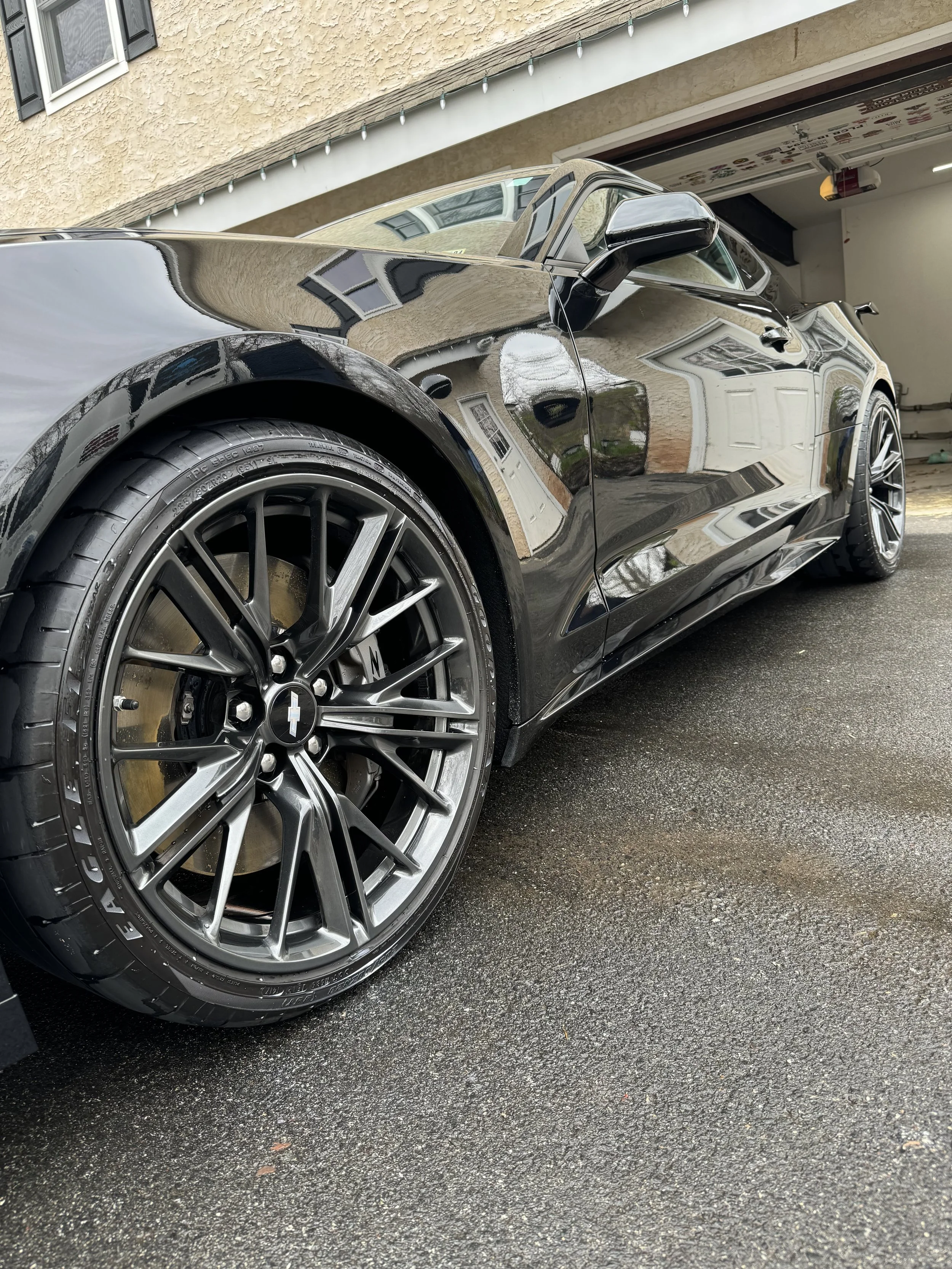 Close-up of a black sports car with shiny, reflective surface, showing the front left wheel with a multi-spoke rim and tire, parked on an asphalt driveway in front of a house with a garage door.