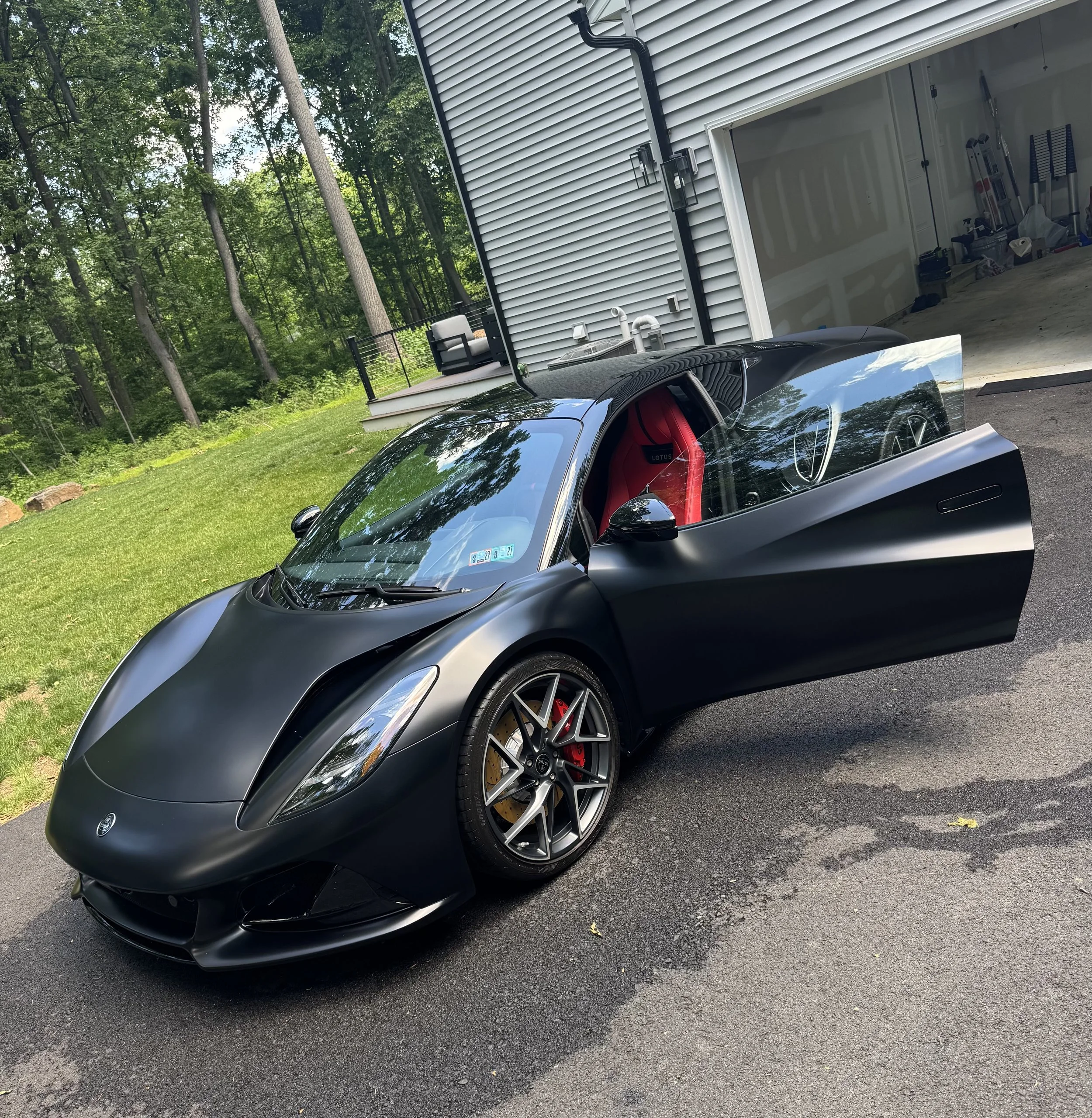 Black sports car parked in driveway beside a garage, with a wooded backyard and a small deck with outdoor furniture visible in the background.