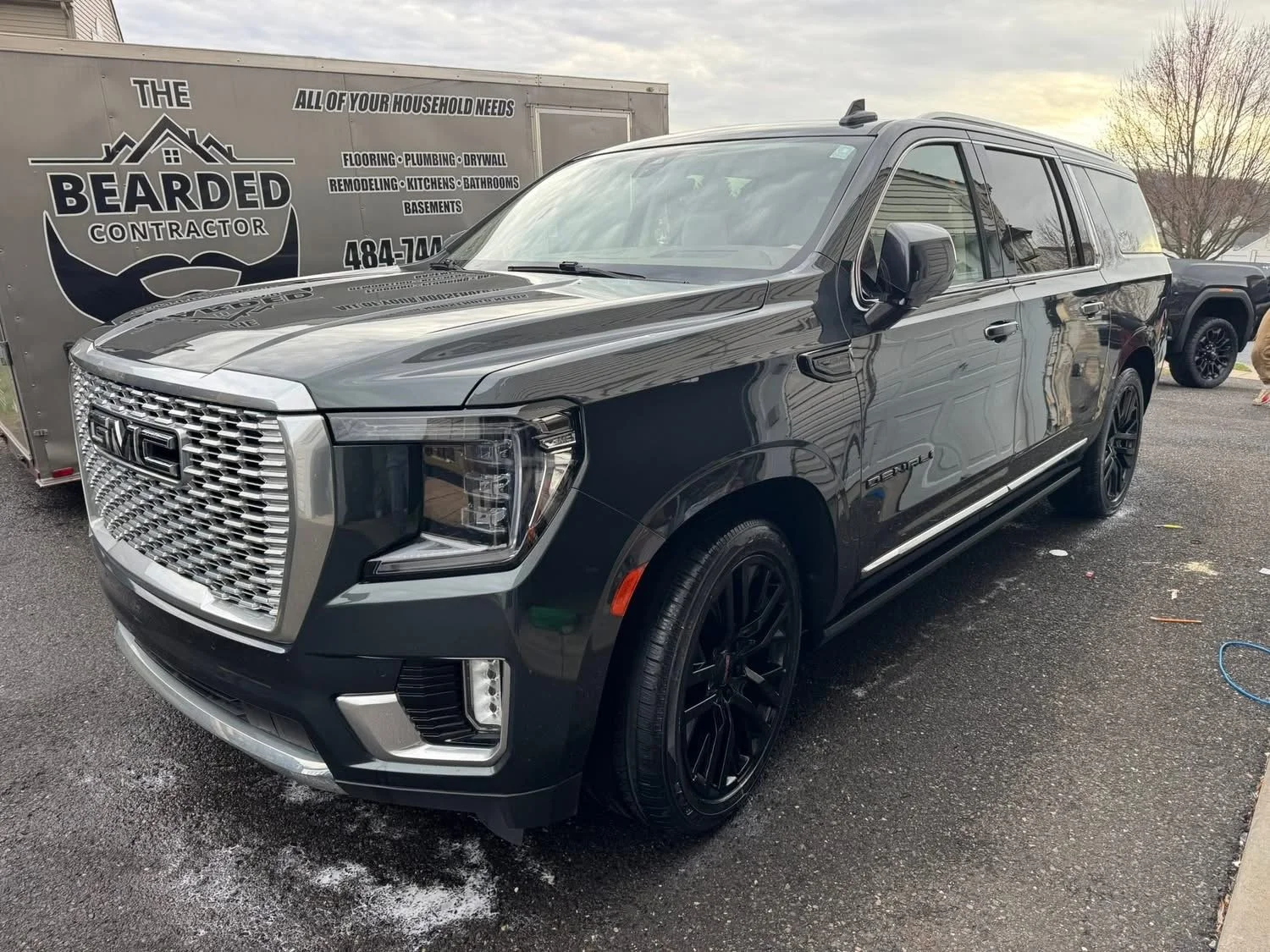 A black GMC SUV parked outdoors on a wet asphalt surface with a bearded contractor trailer in the background and overcast sky.