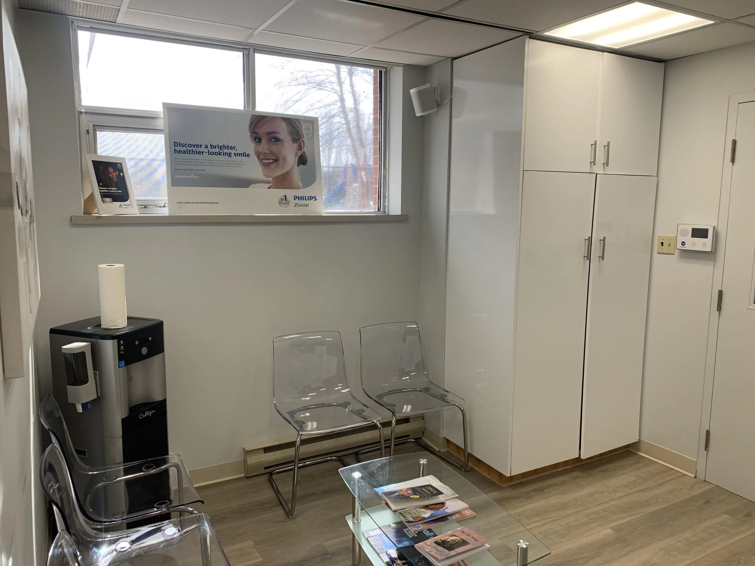 A waiting room with transparent chairs, a glass coffee table with magazines, a water dispenser, white cabinets, a large window with outdoor trees, and a promotional poster for Philips teeth whitening on the window sill.