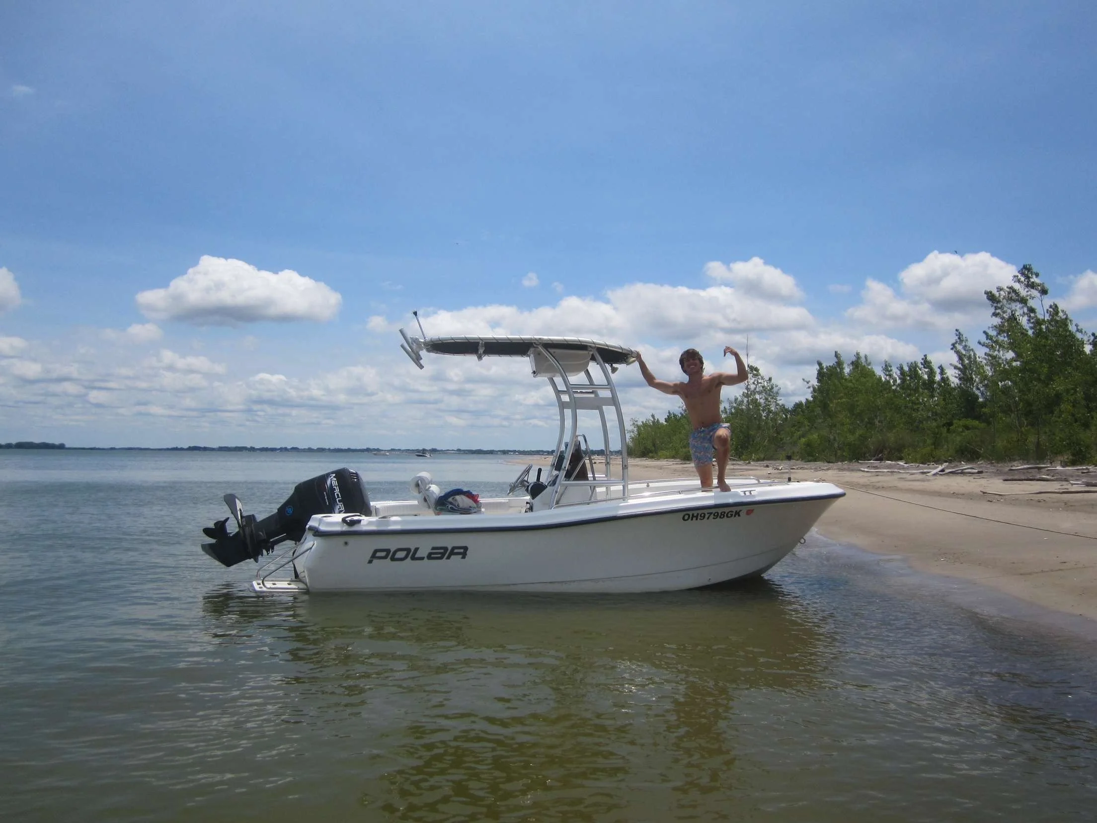 A man kneeling on the bow of a white boat at a lakeshore, flexing his arm.