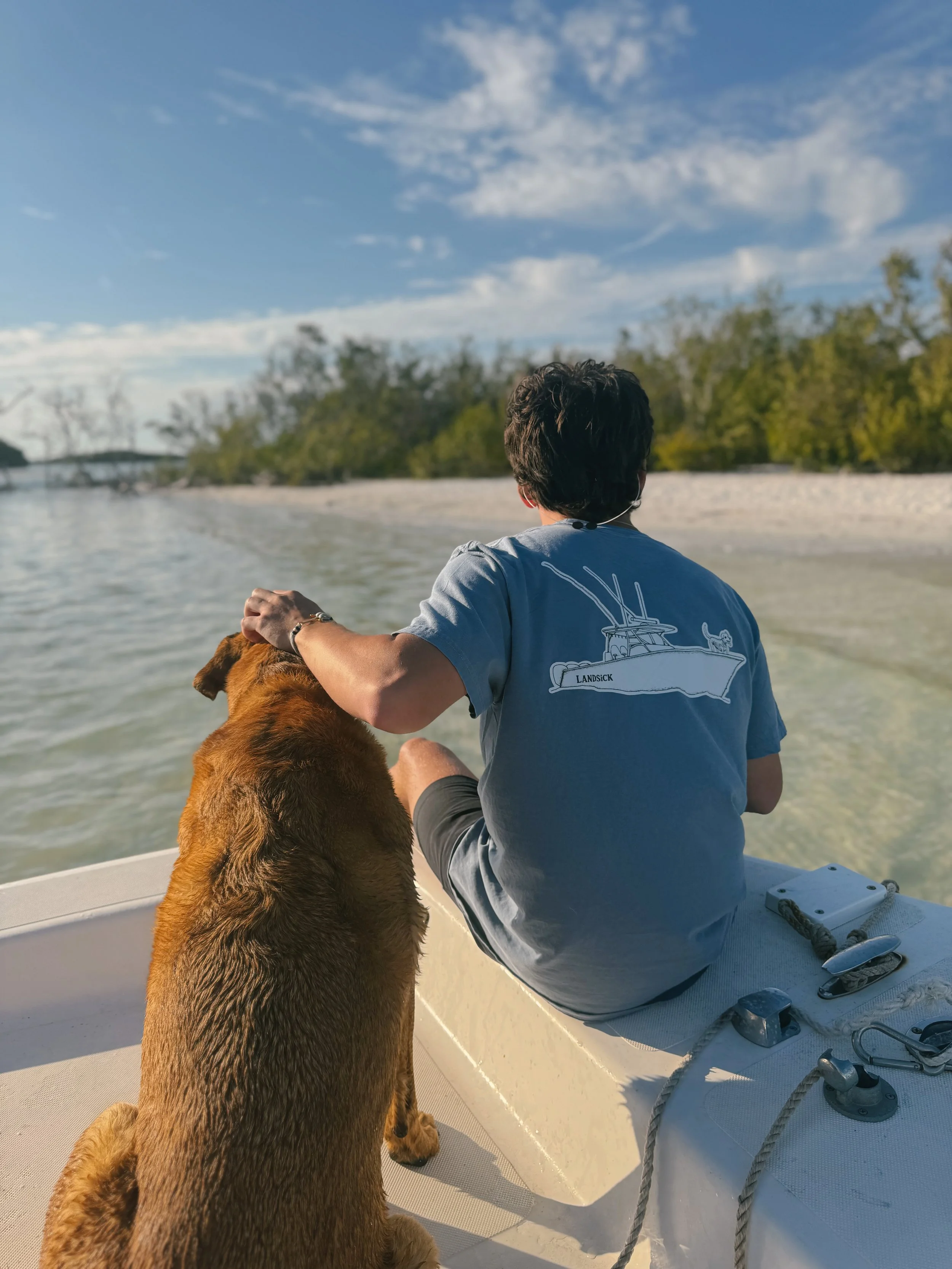 A man and a dog sitting on a boat, looking at a shoreline with trees under a blue sky with clouds.