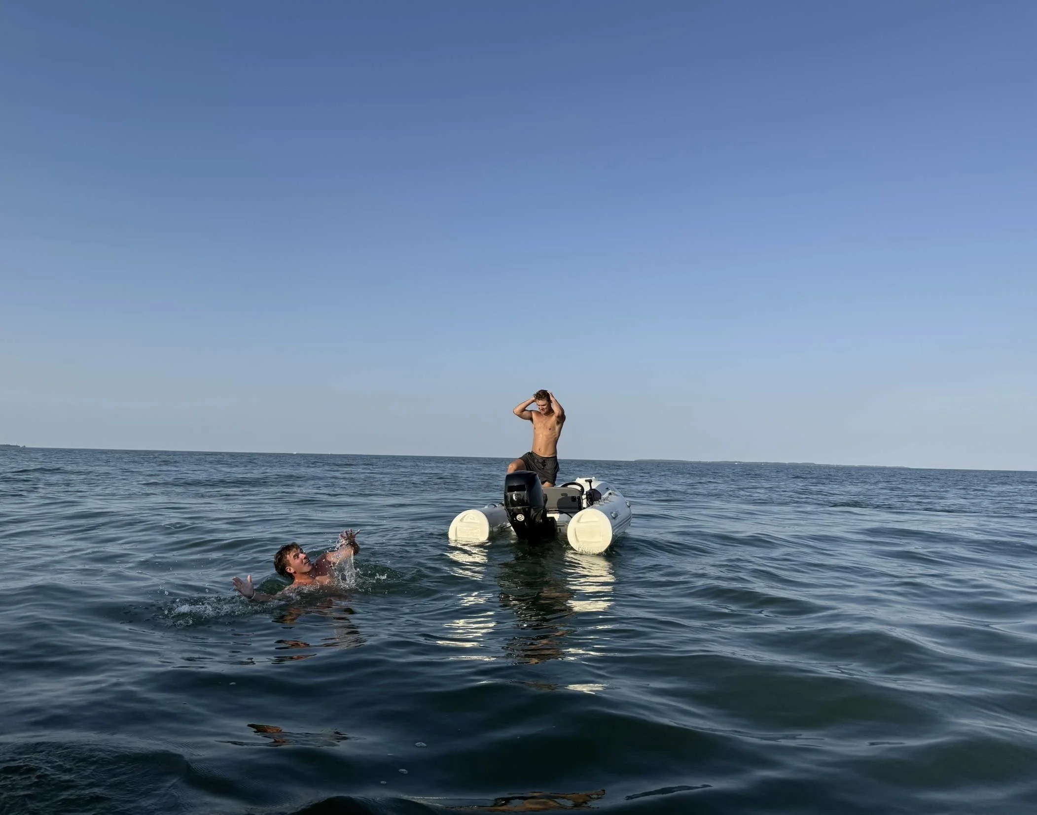 Two people on open water, one standing on a small boat and one swimming nearby.