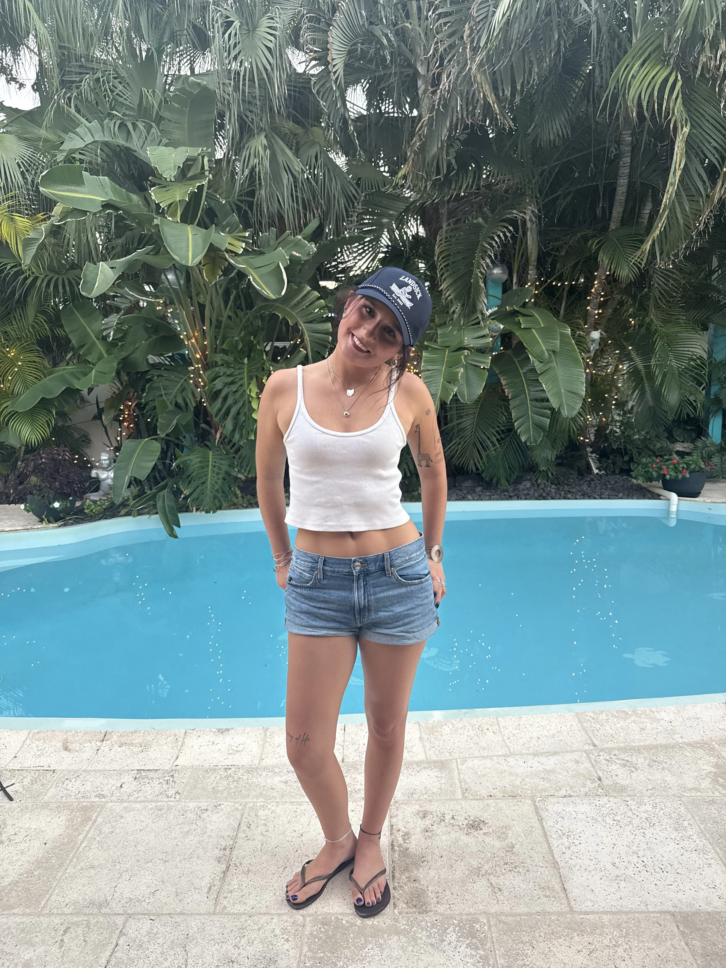 A woman with long dark hair, wearing a baseball cap that says "Landsick" and a white tank top, sitting outdoors at a waterfront restaurant or bar with string lights overhead and boats in the water behind her.
